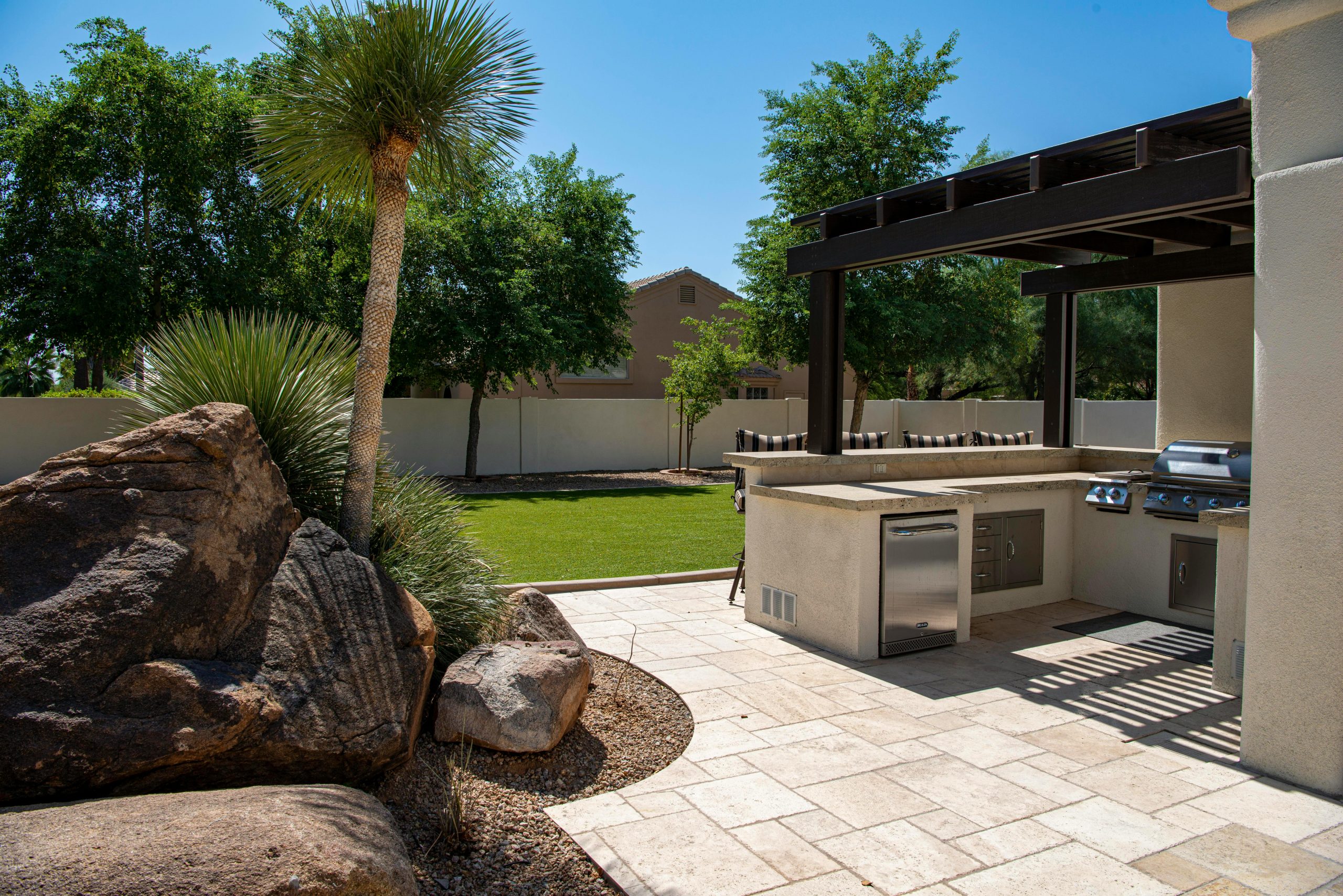 Stylish outdoor kitchen on a sunny patio with lush landscaping and clear blue sky.