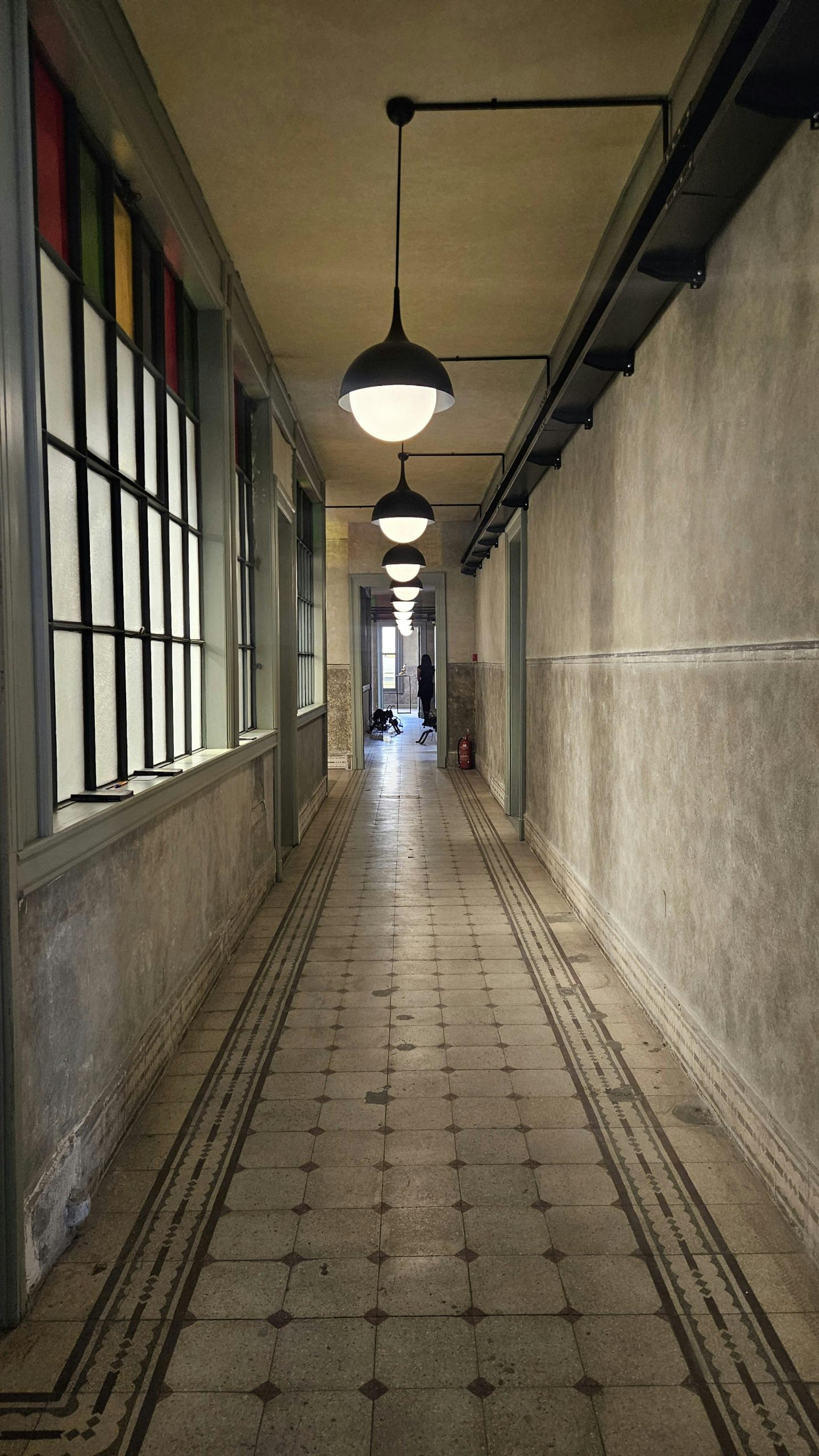 Long vintage hallway with hanging lights, tiled floor, and large windows.