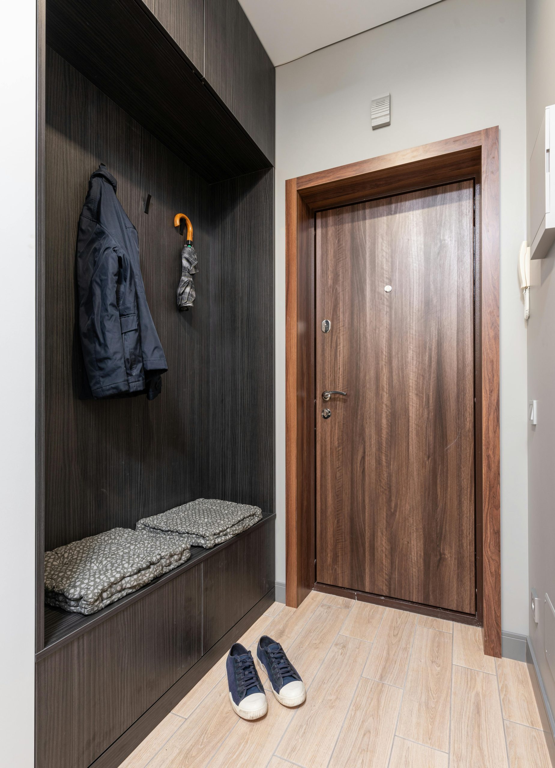 Modern light apartment interior with wooden wardrobe with jacket near umbrella and textile on shelf near sneakers and door