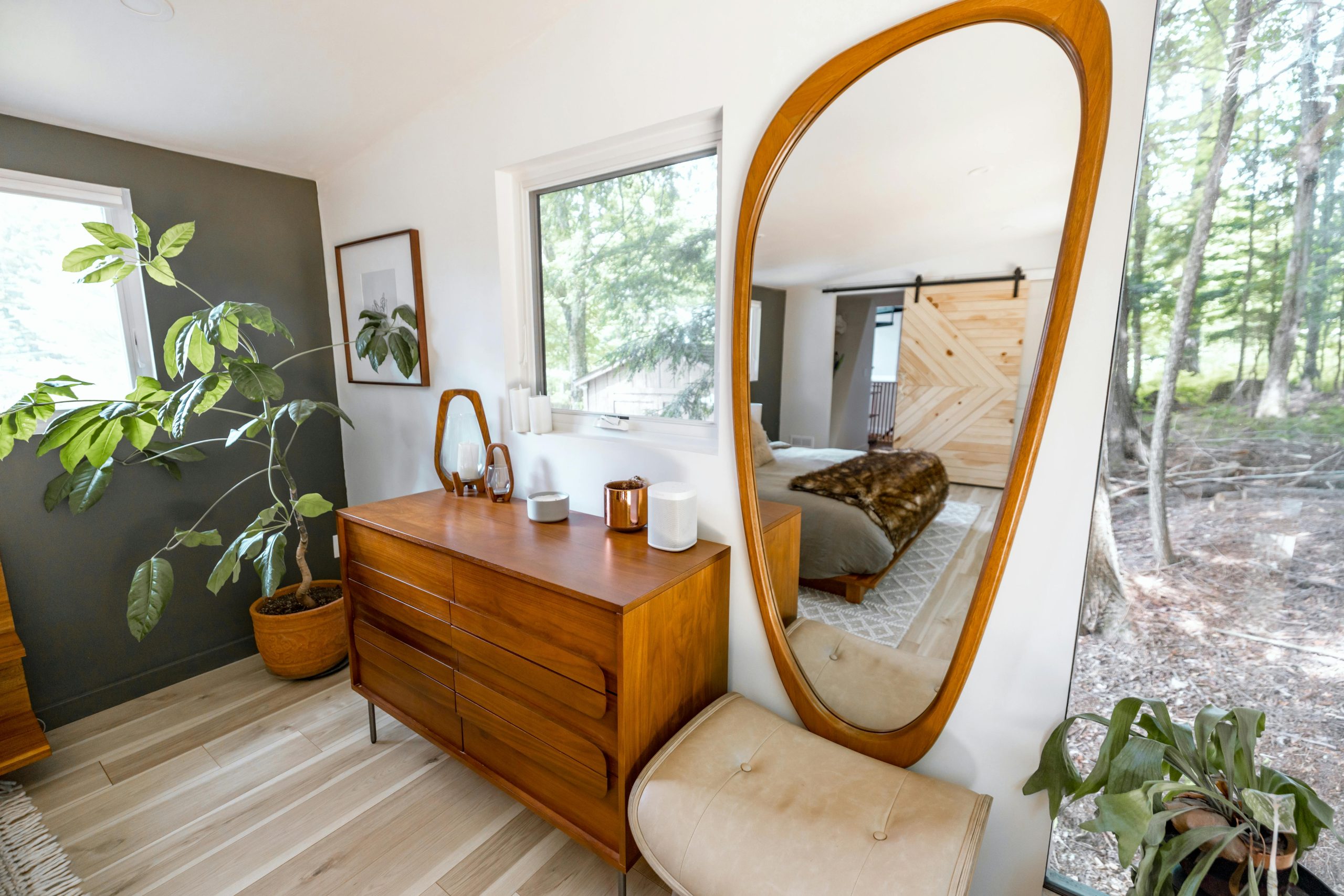 A stylish bedroom interior featuring a wooden dresser, large mirror, and indoor plants under natural light.