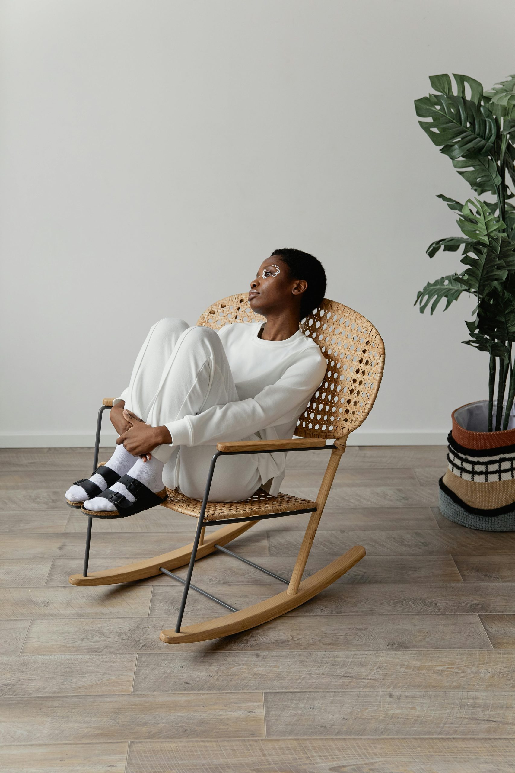 A thoughtful woman sits in a modern rocking chair indoors with plants nearby.