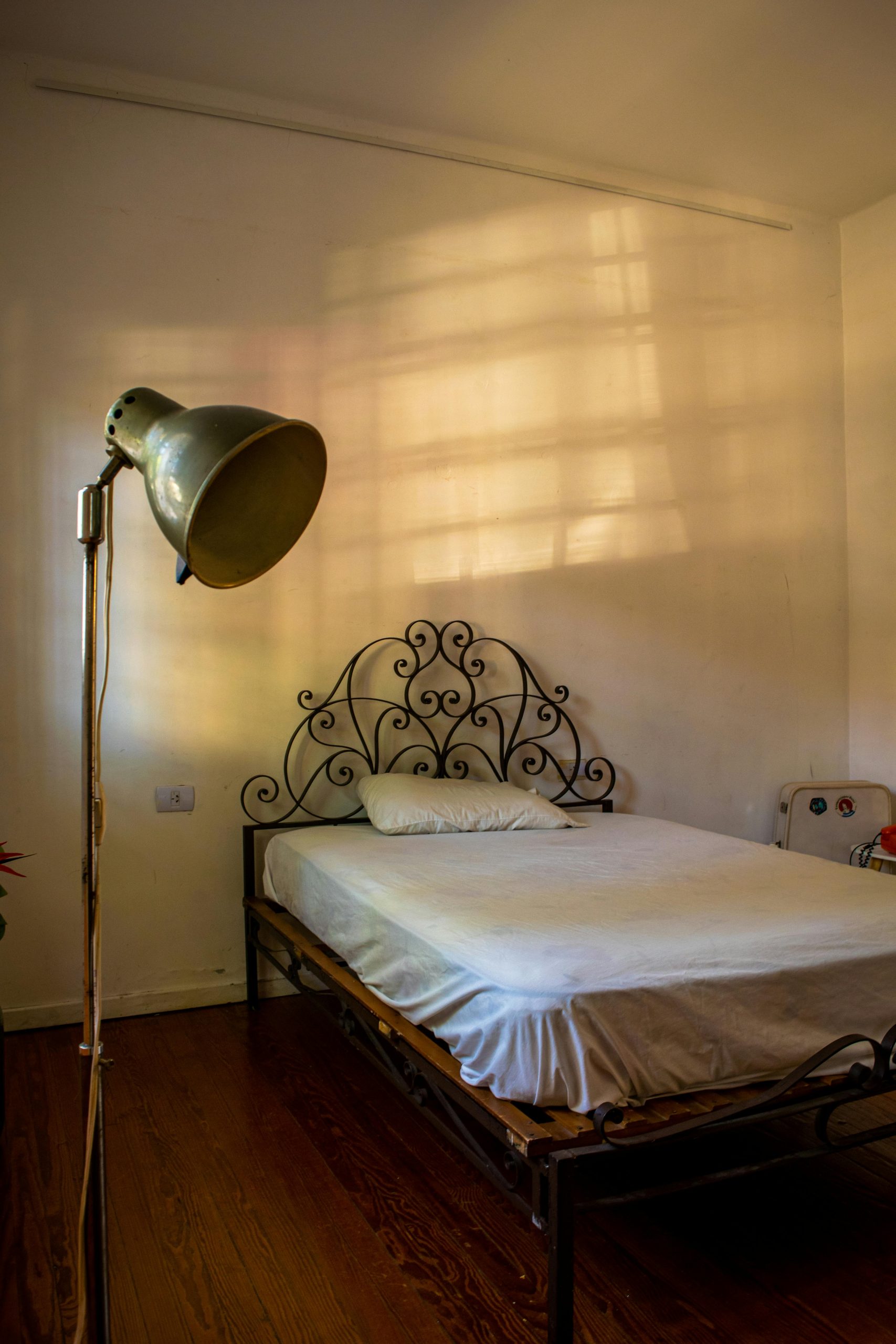 A vintage bedroom with soft natural light casting shadows on the wall, featuring an ornate metal bed and floor lamp.