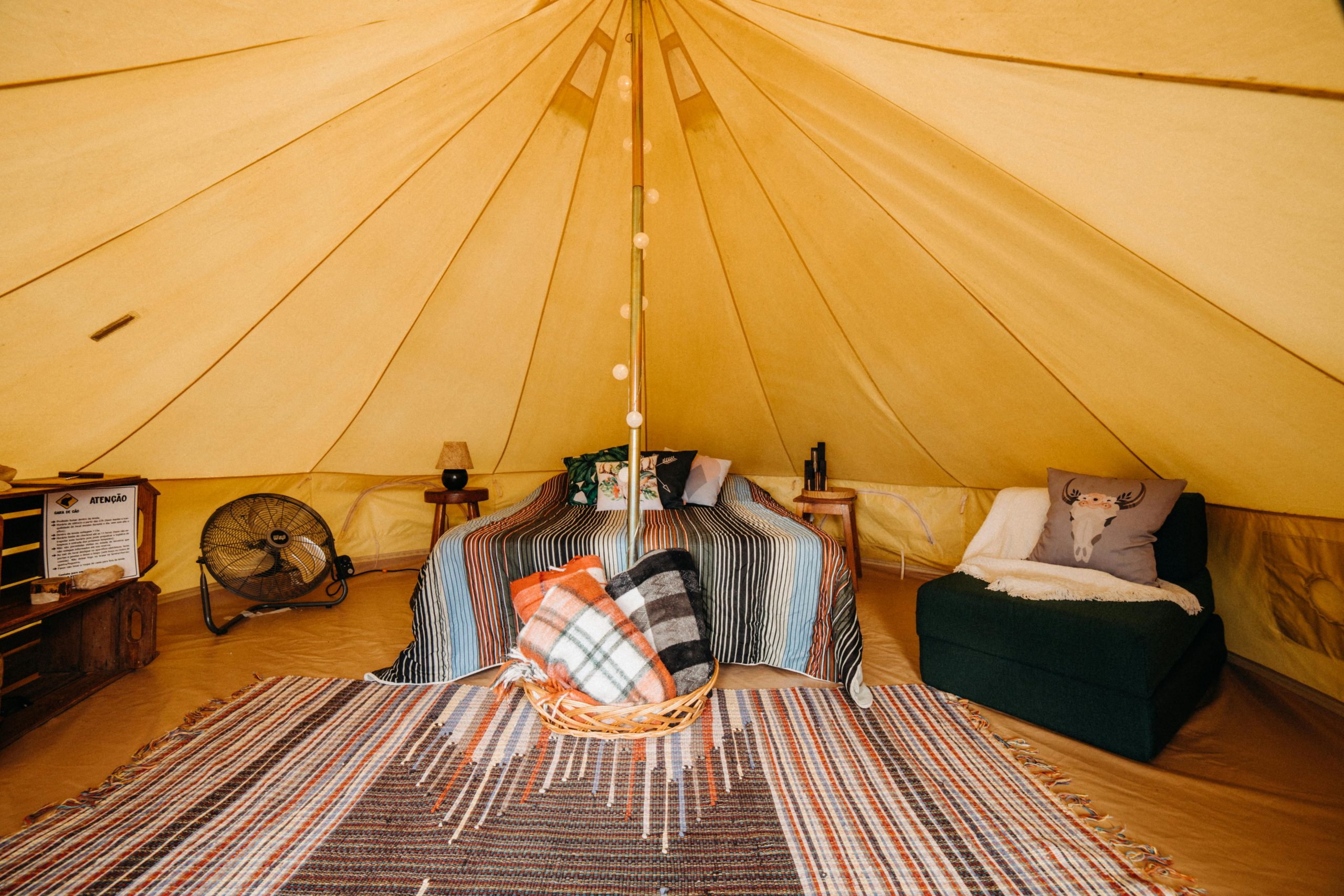 Bed with colorful blanket near comfortable armchair with cushions placed in yellow spacious tent with colorful carpet and decorated pole