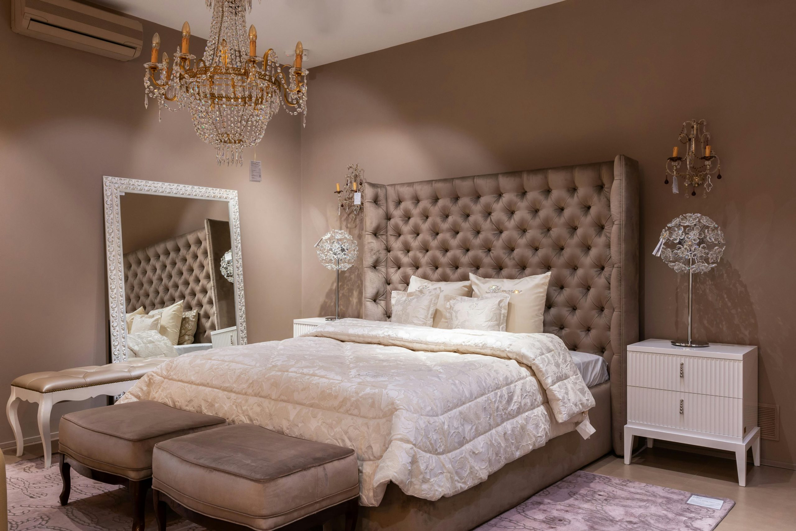 Comfortable bed with bedclothes and upholstered headboard placed at wall near mirror and bedside table in spacious bedroom with benches