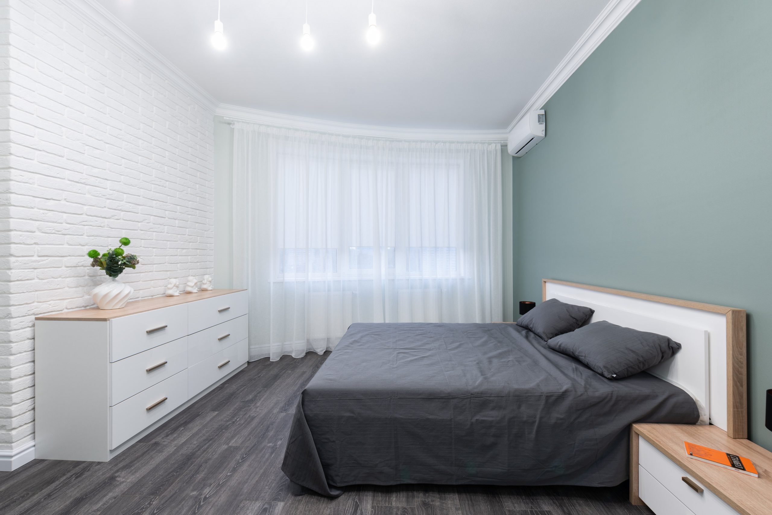 Contemporary bedroom interior with bed against table and chest of drawers on parquet under shiny light bulbs