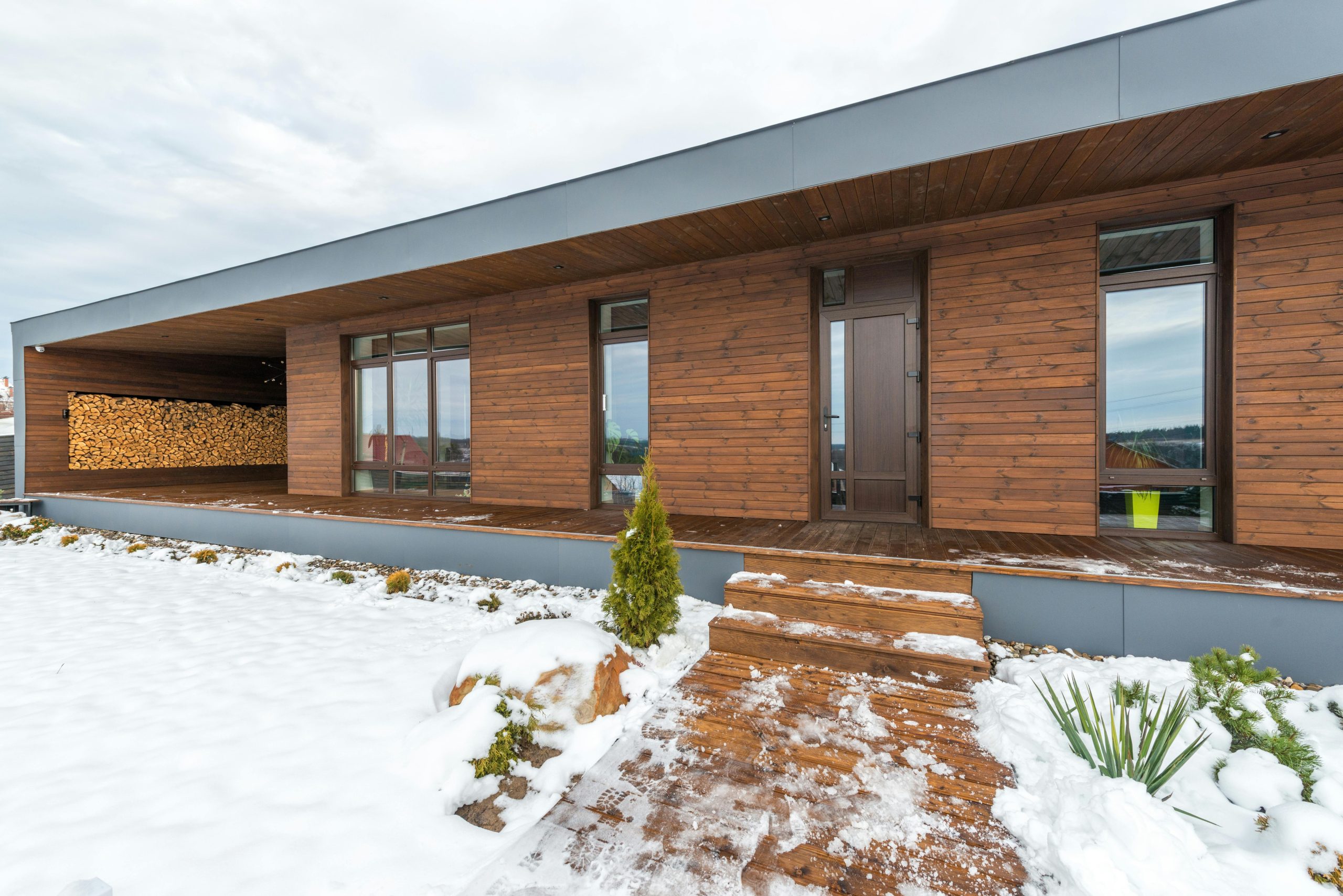 Contemporary wooden house with snow-covered yard and stacked wood for winter.