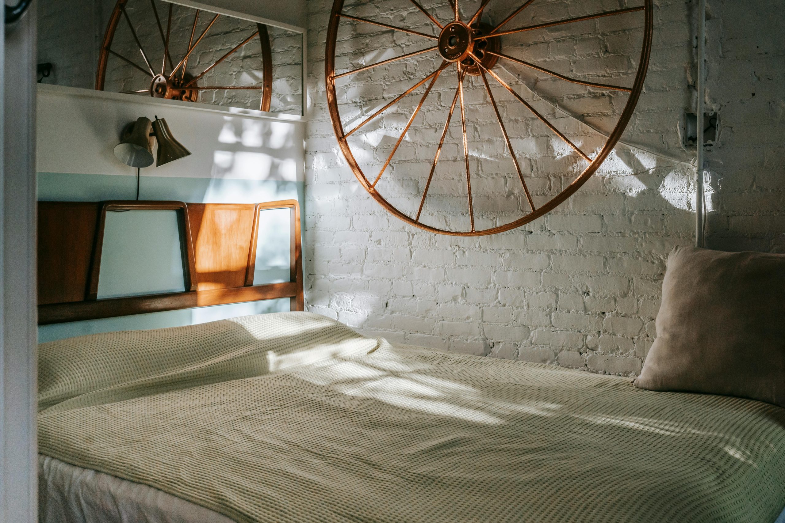Cozy bed under lamps and old big wheel on brick wall in sunlight at home
