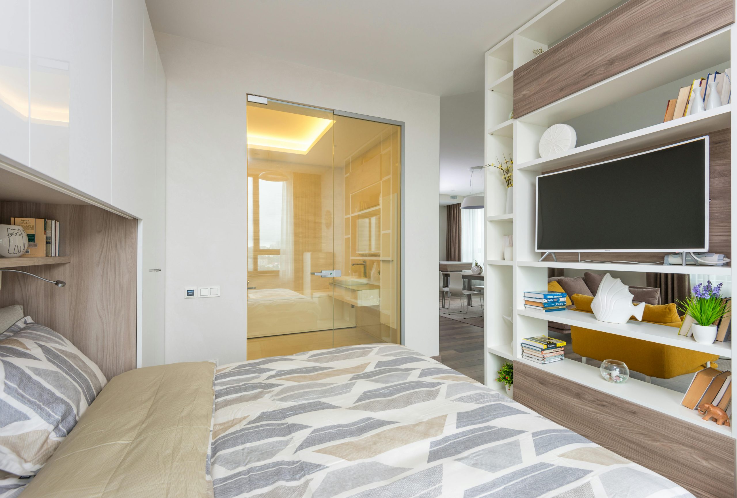 Interior of light cozy bedroom with open plan living room behind shelf partition with TV and opened glass doors to bathroom