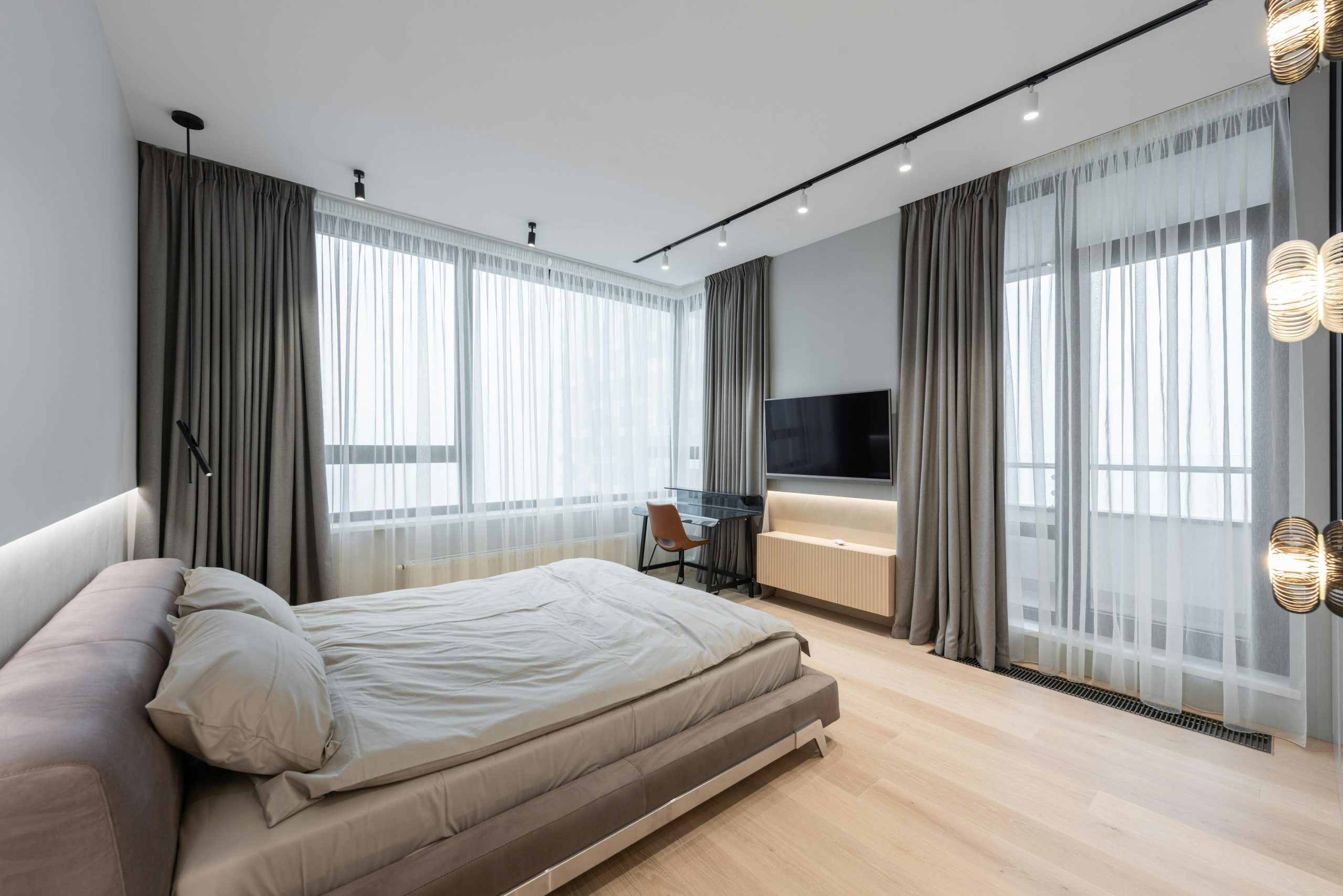 Interior of modern light bedroom with bed next to TV with curtains on windows and table with chair