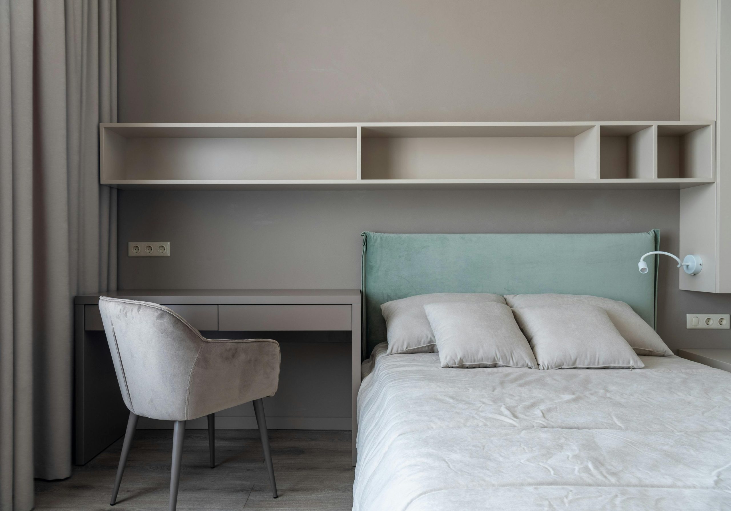 Interior of modern light bedroom with pillows on bed near table with chair under shelves next to curtains