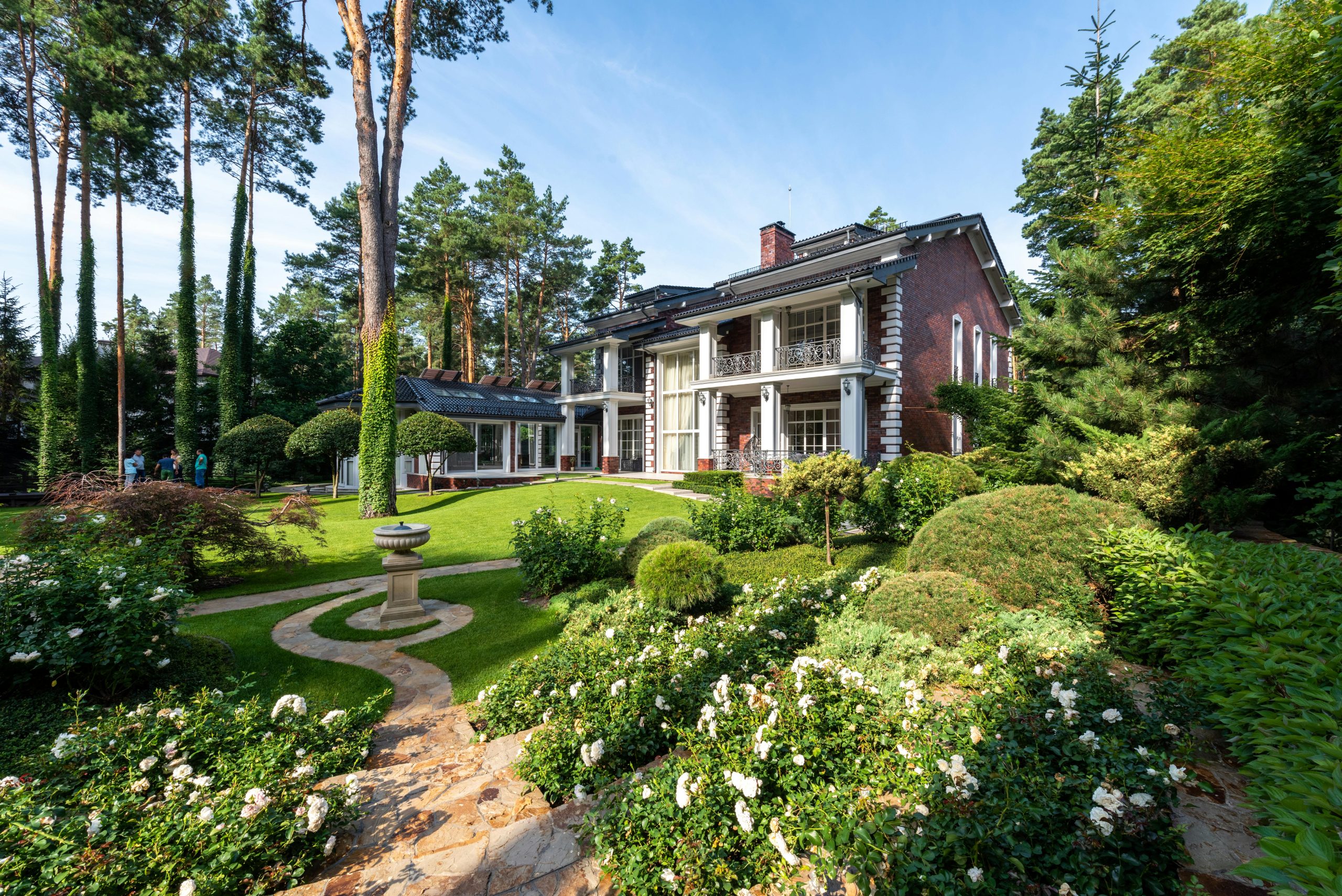 Modern house near yard with green grass and trees near plants and path in sunny summer day under blue sky