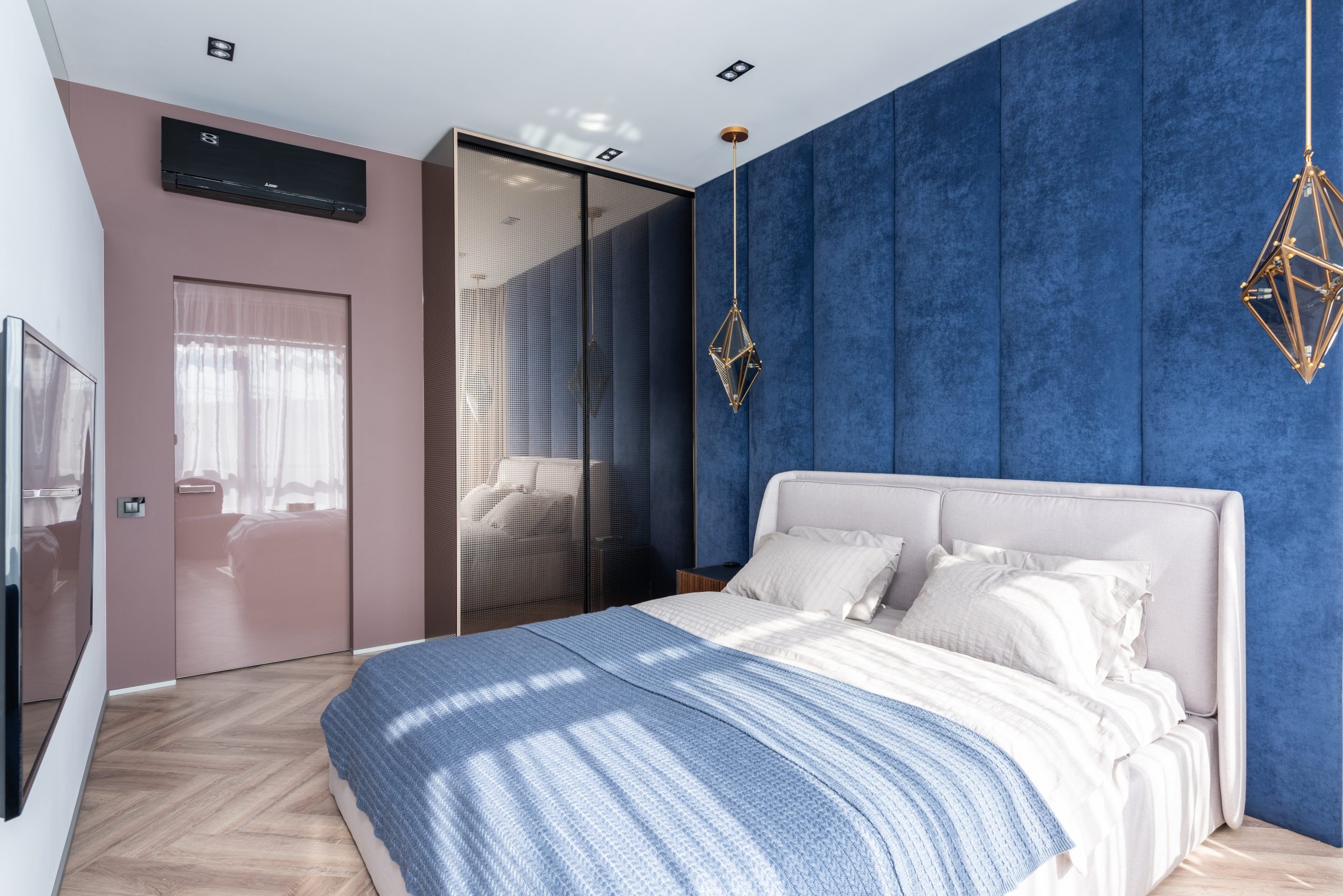 Soft bed with blanket placed against white wall with TV in modern bedroom with wardrobe and door near blue wall