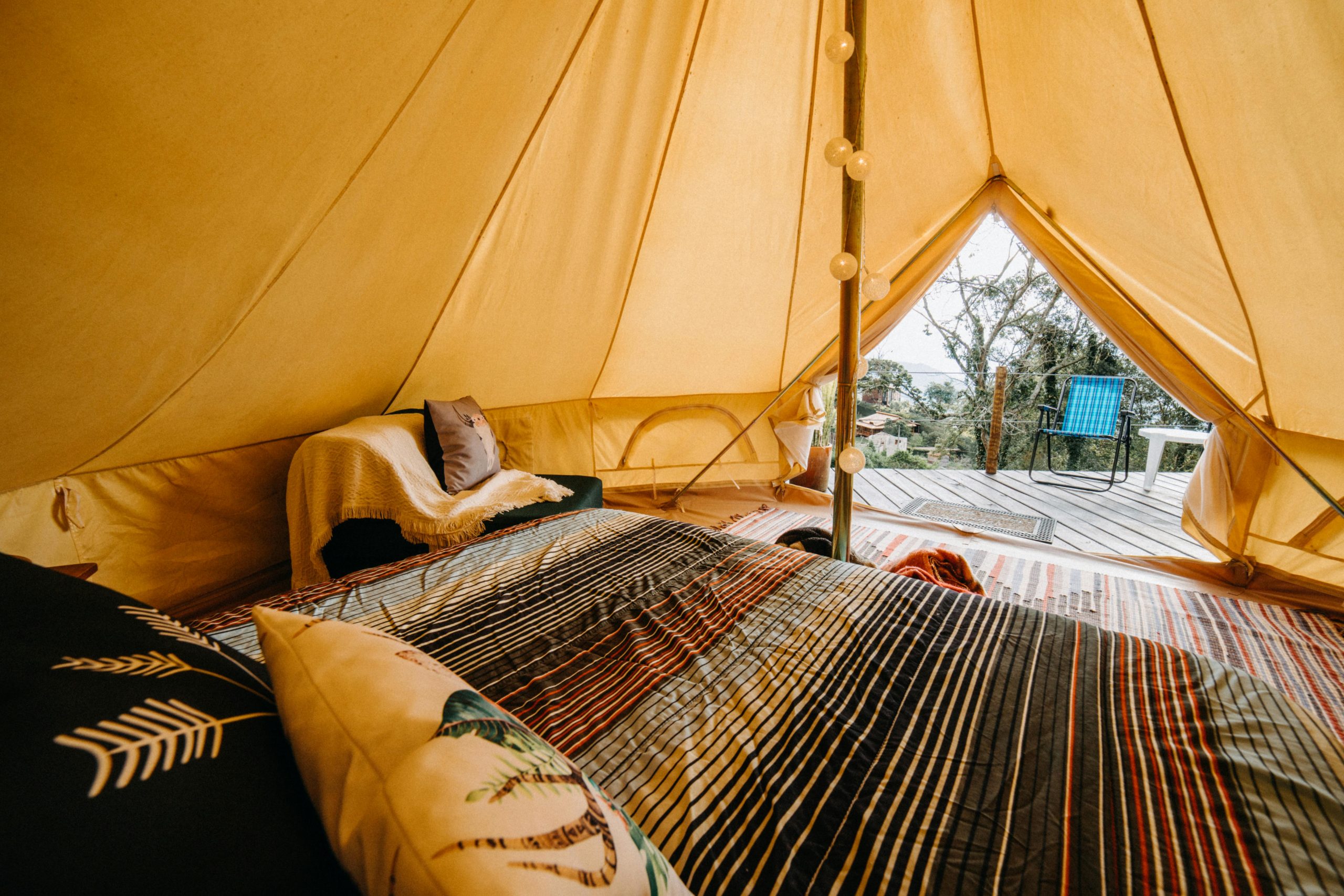 Soft bed with colorful blanket and cushions placed in tent with decorated pole and doorway in rural area of countryside