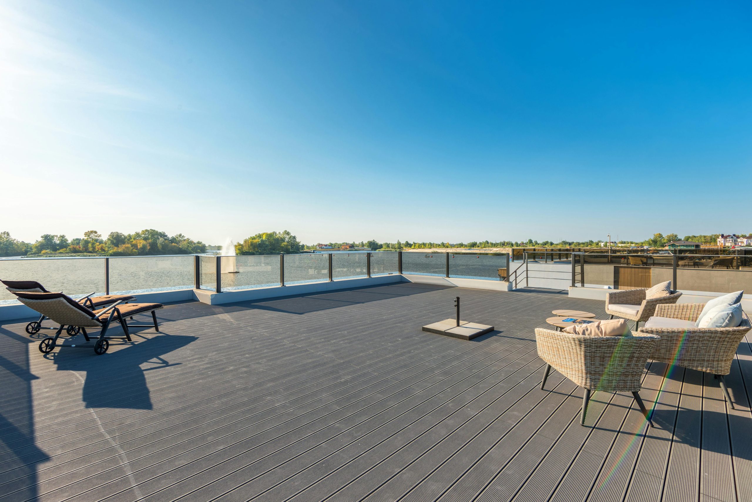 Spacious, sunny waterfront deck with modern wicker seating and lounge chairs overlooking a serene lake.
