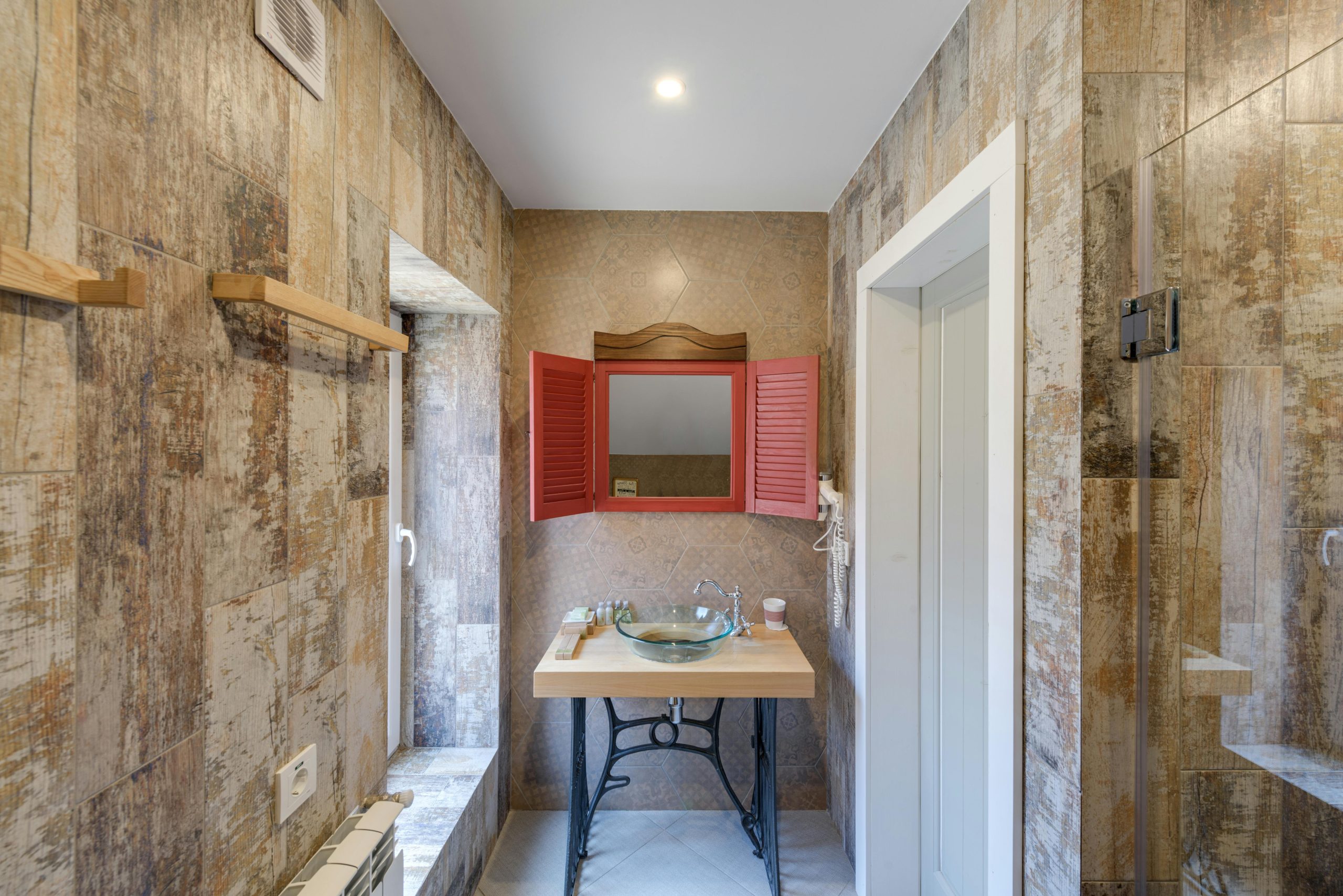 A stylish bathroom with rustic elements featuring a wooden vanity, glass sink, and red shutters.