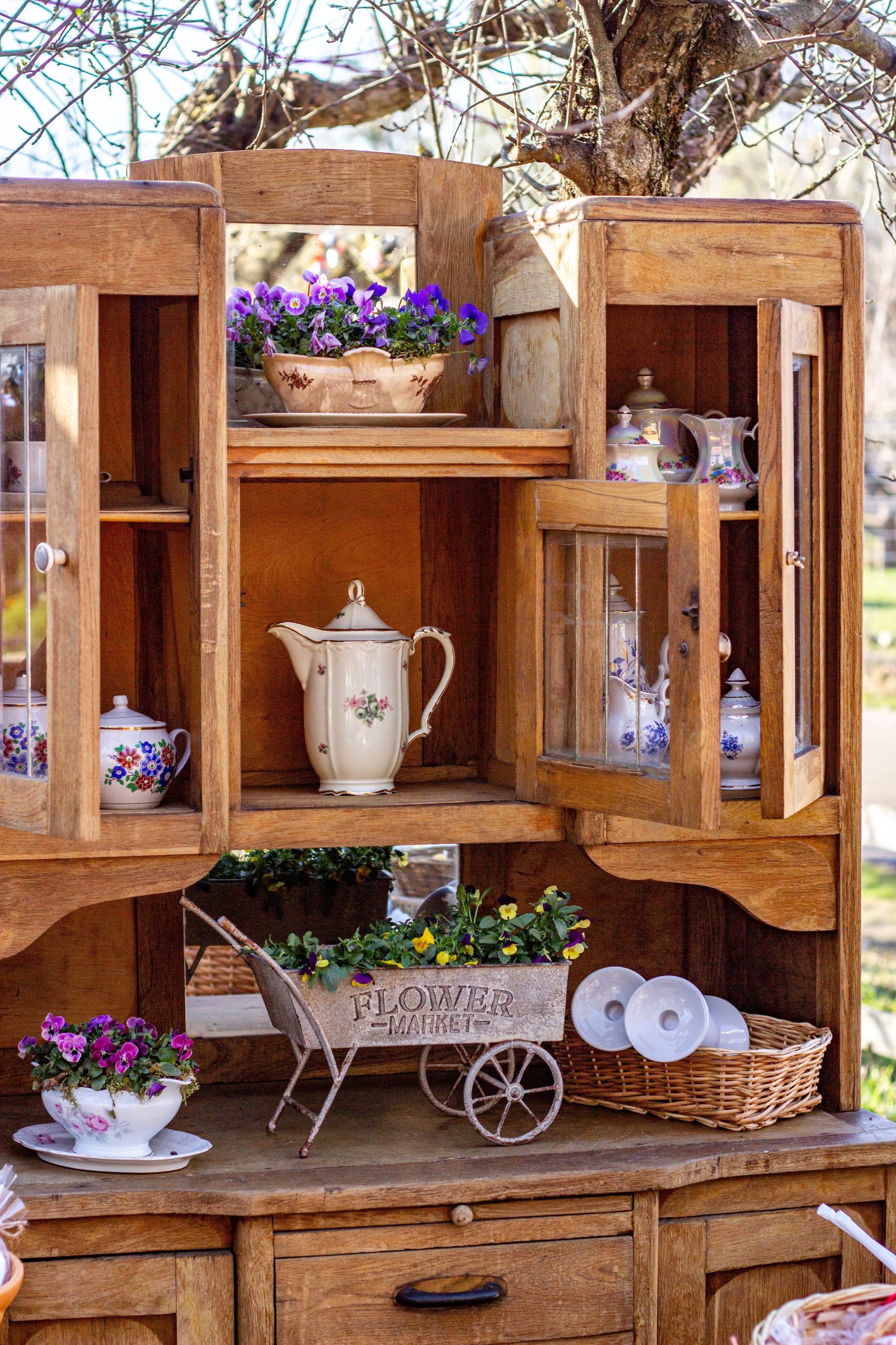 Charming vintage wooden cabinet with teacups and decorative flowers under a sunny tree.