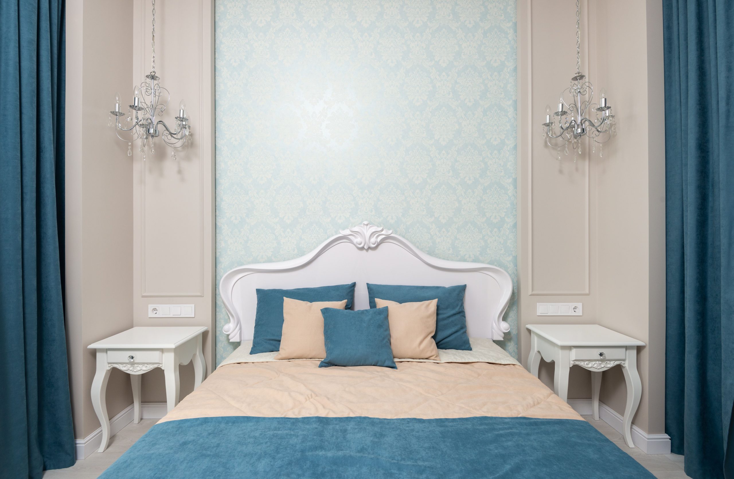Comfortable bed with blue and beige bedclothes between bedside tables at wall with wallpaper and hanging lamps in bedroom with classic design