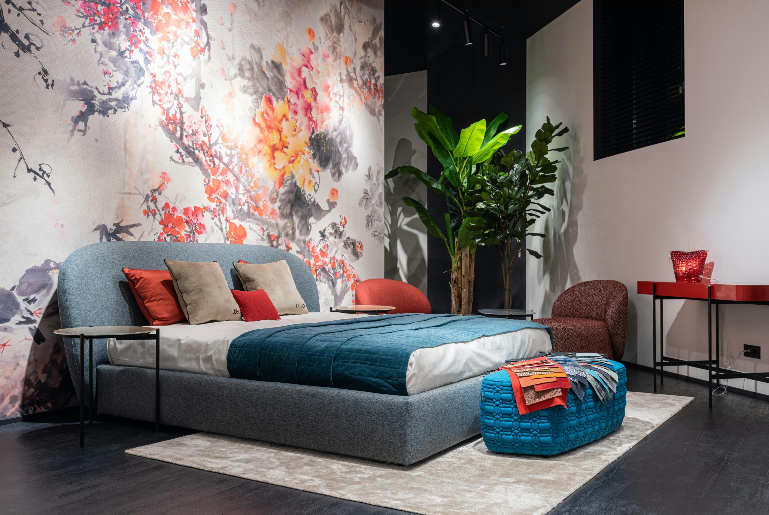 Comfortable bed with colorful cushions and blue blanket placed at painted wall in modern bedroom with armchairs and potted plant
