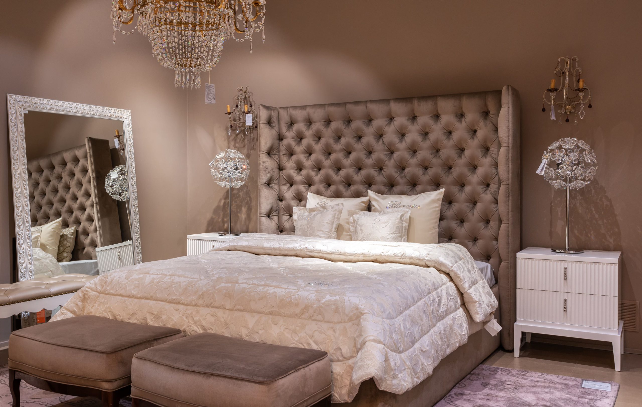 Comfortable bed with cushions and blanket placed at wall near mirror in spacious bedroom with soft benches and chandelier in apartment