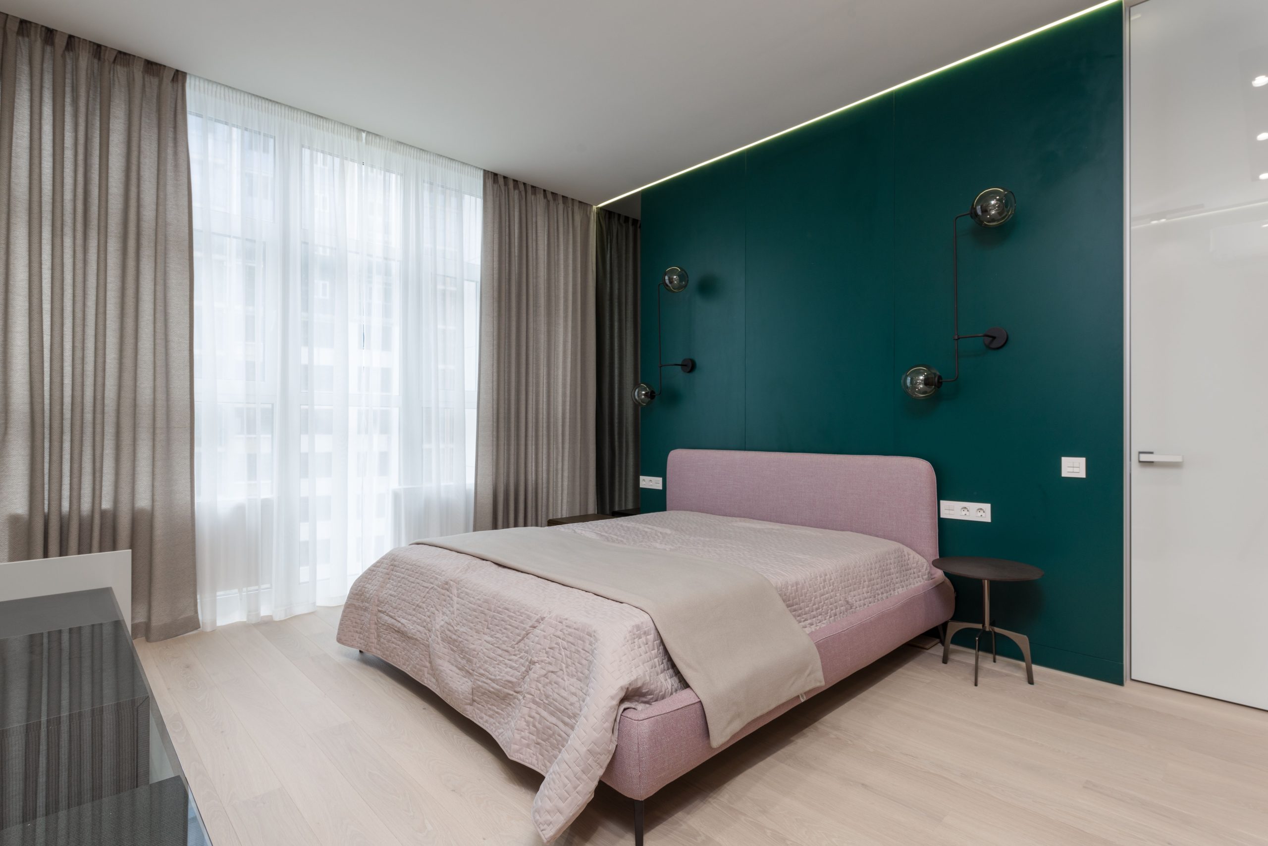 Comfortable bed with small round bedside tables placed in modern room with chiffon curtains and green wall decorated with creative lamps