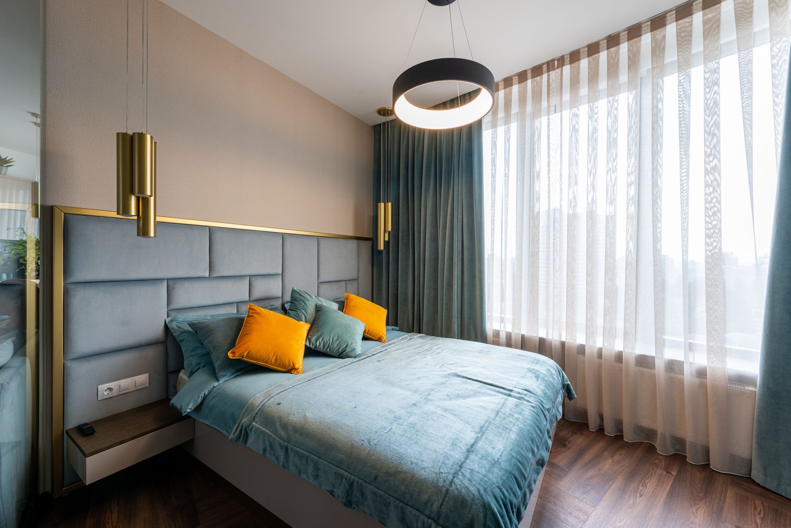 Cozy bed with turquoise blanket and colorful cushions placed near window with curtains in modern bedroom with hanging lamps at home
