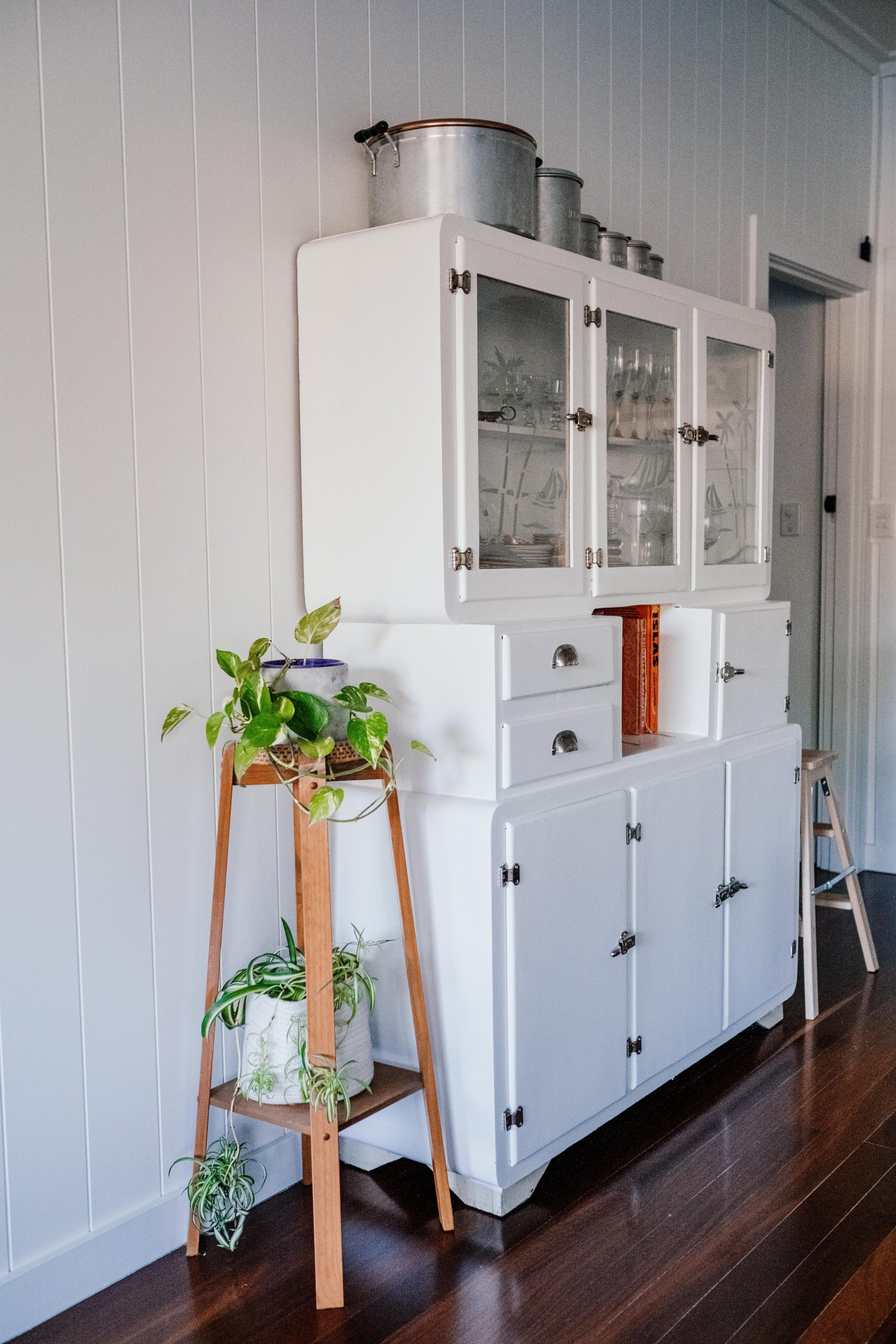 Elegant white vintage kitchen cabinet with glass doors and potted plants in a serene home setting.