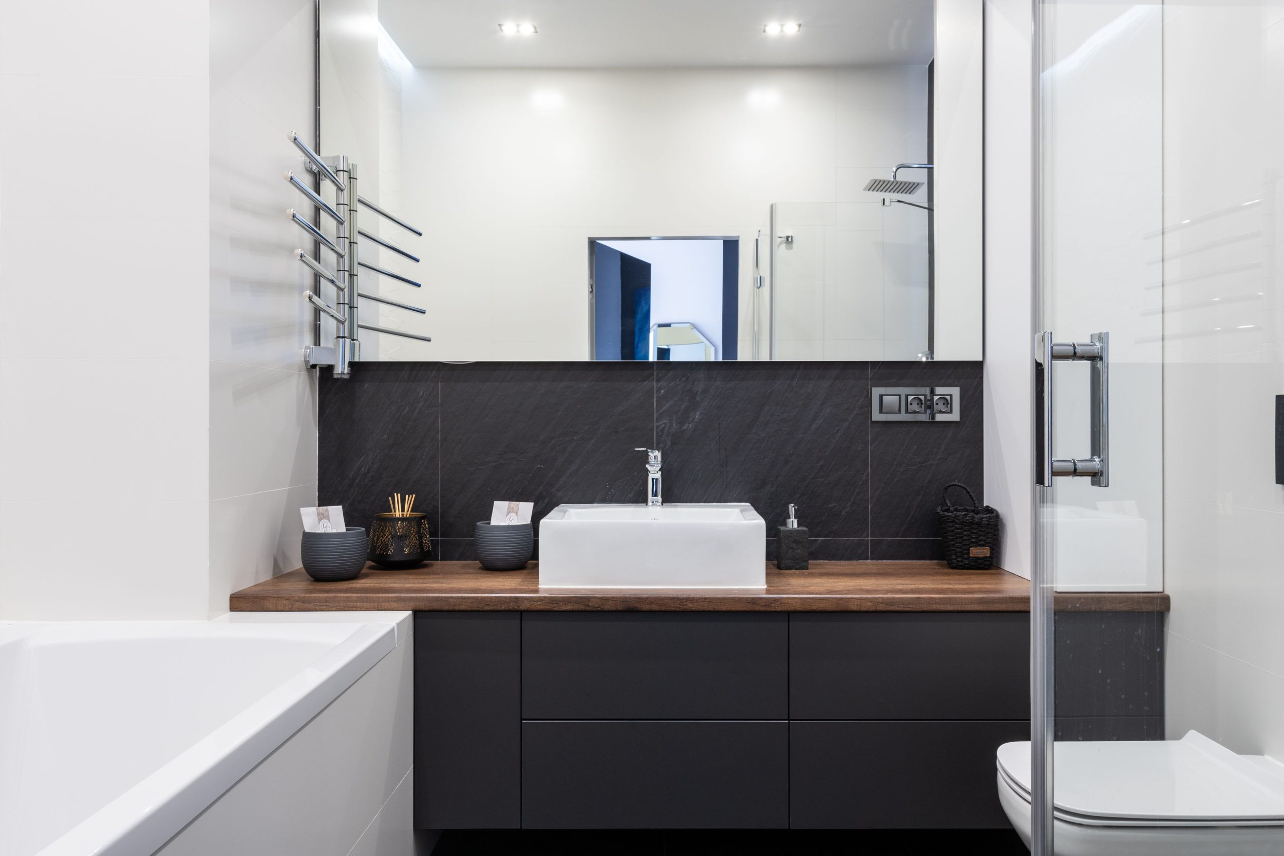 Interior of contemporary bathroom with shower cabin white tub and toilet near black cupboard with sink placed in room with mirror
