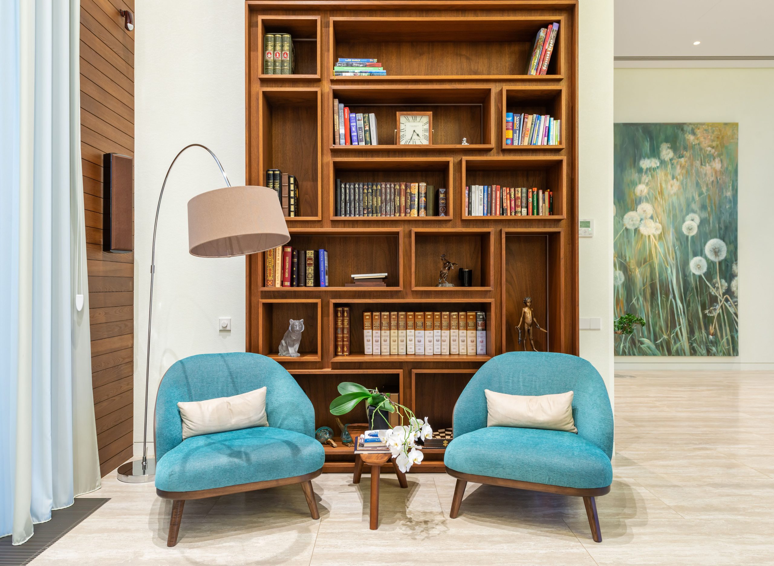 Interior of living room with cozy soft blue armchairs near lamp and books placed on shelves of wooden bookcase