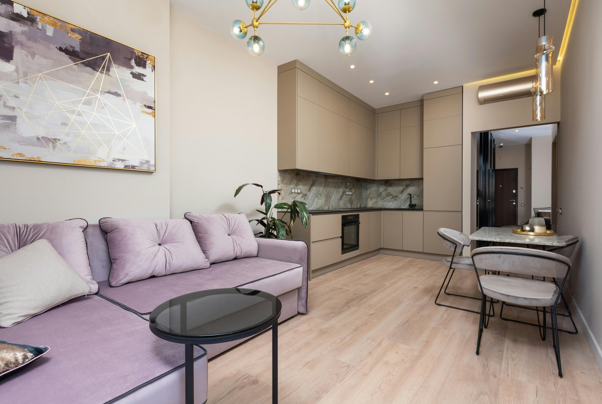 Interior of modern light studio with couch with pillows near table and chairs next to open kitchen with cabinets near painting on wall