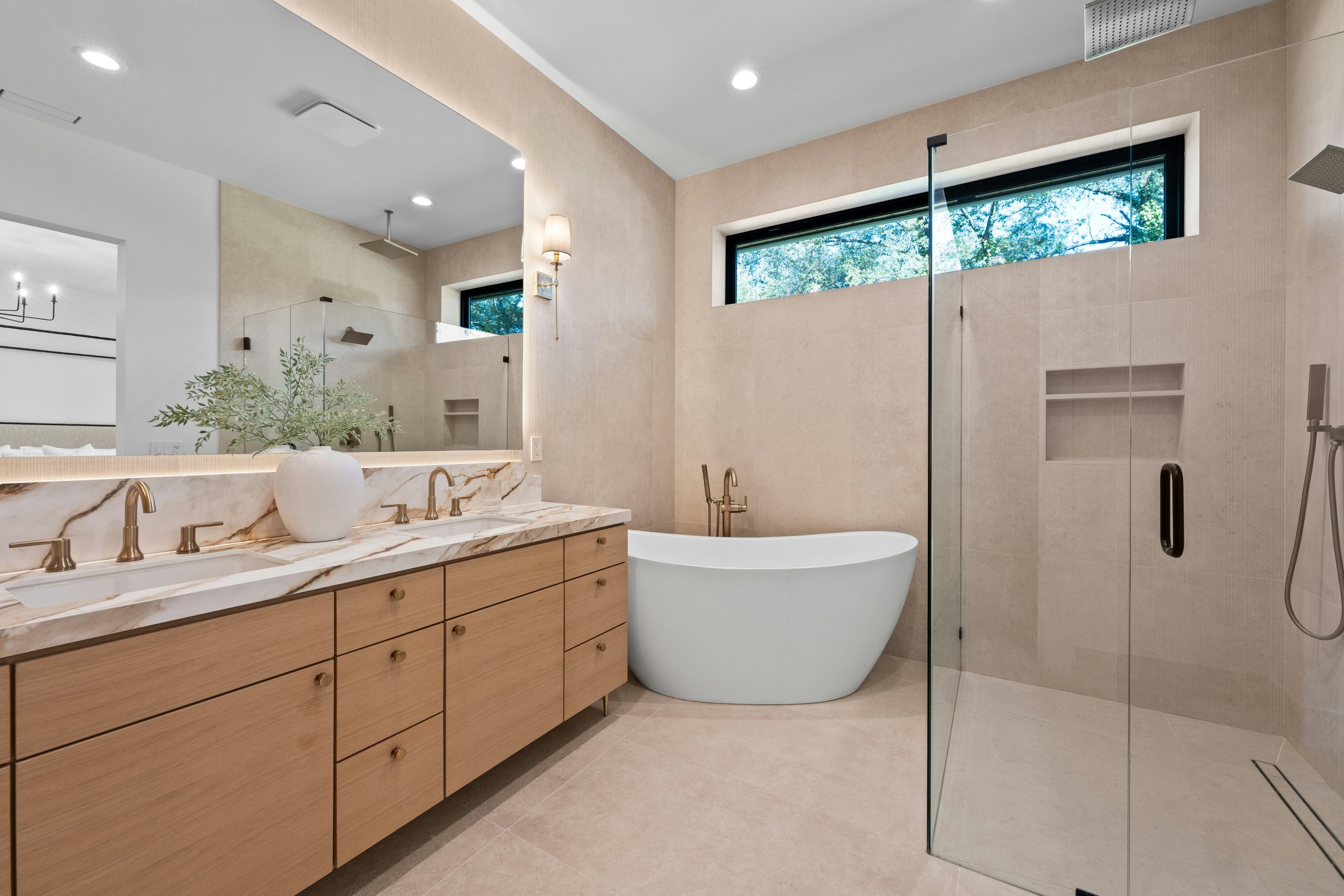 Luxury modern bathroom with oak double vanity, marble countertop, and freestanding soaking tub. Contemporary custom home featuring a glass walk-in shower, brass fixtures, and warm neutral tile. High-e
