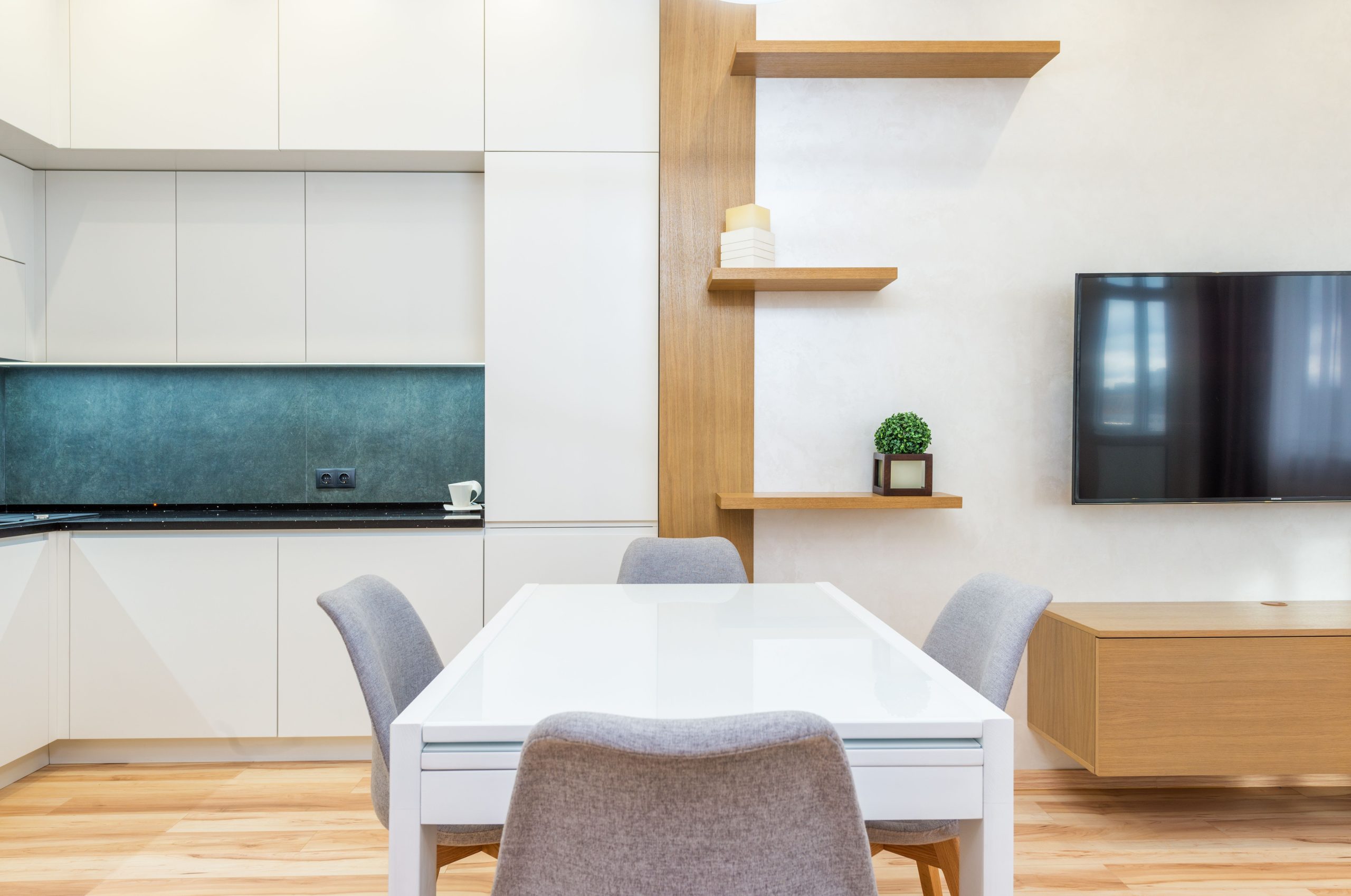 Modern apartment interior with table with chairs next to shelves and open kitchen with cabinets near TV on wall