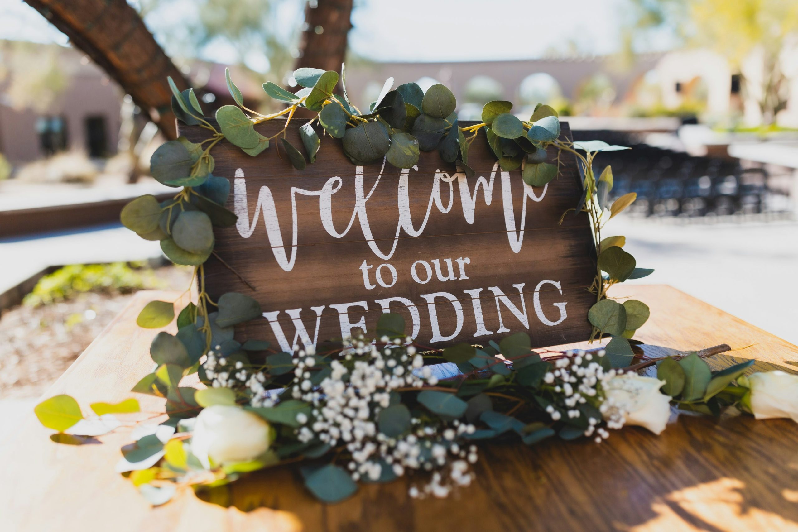 Rustic wooden 'Welcome to our Wedding' sign decorated with eucalyptus garland and baby's breath flowers. Outdoor wedding ceremony entrance decor with hand-lettered calligraphy on dark stained wood.