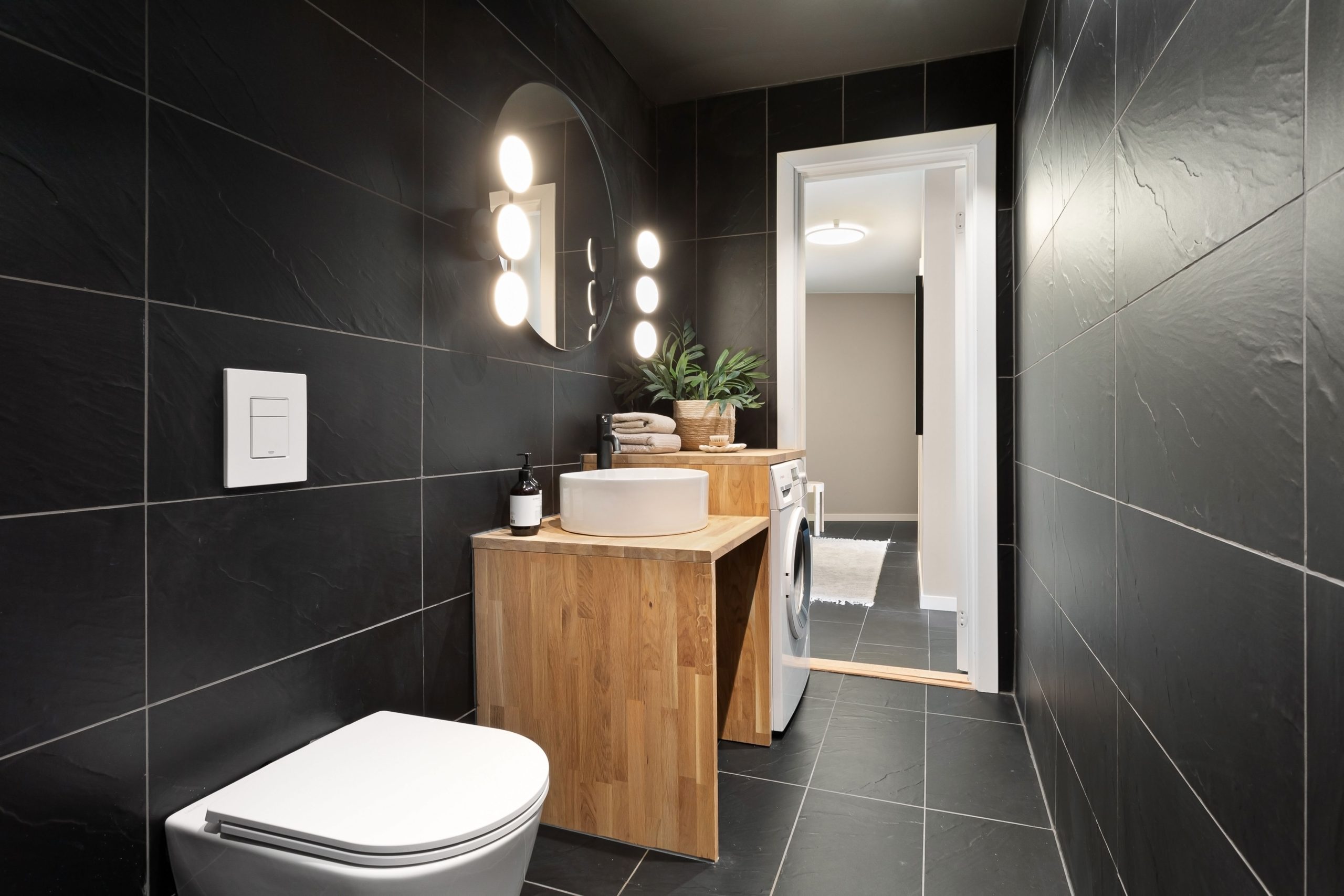 Sleek bathroom interior featuring dark tiles, wooden vanity, and mirror lighting.