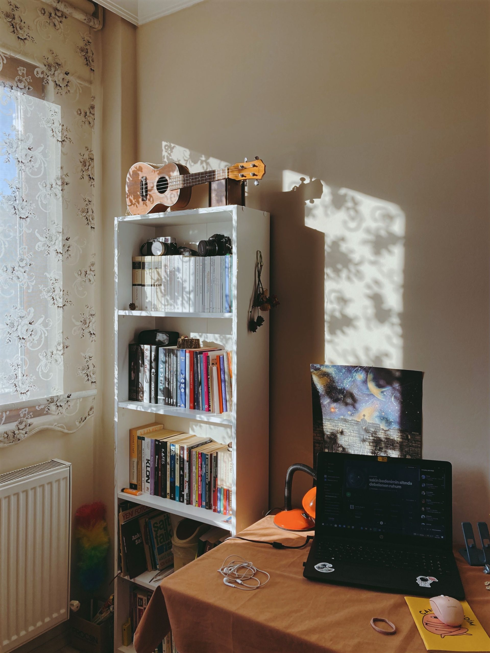 Sunlit study room featuring a bookshelf, laptop, and musical instrument for a cozy ambiance.