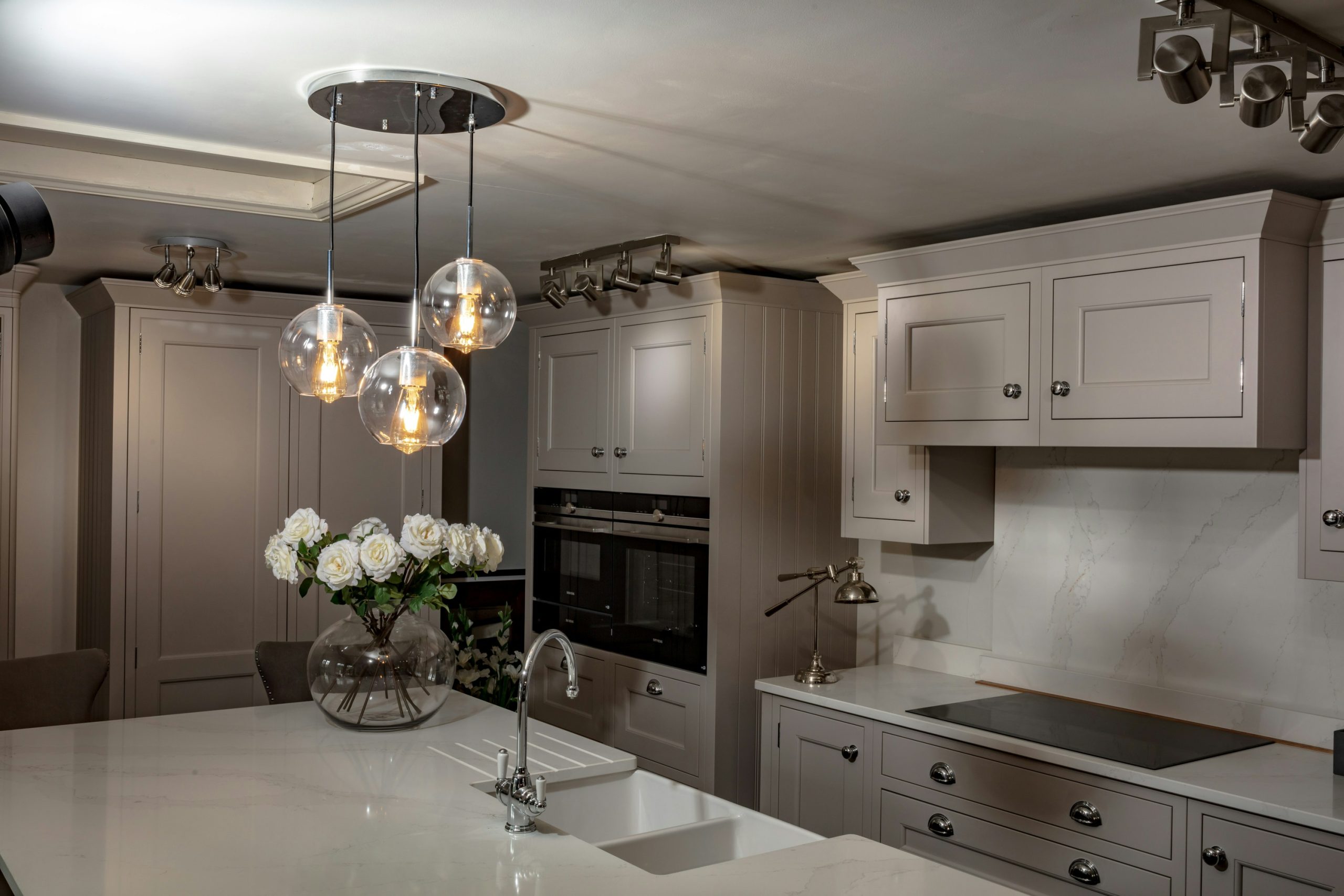 This kitchen interior features a modern, elegant design with light grey cabinetry and sleek, integrated appliances. The focal point is a large island with a white countertop, complete with a sink and
