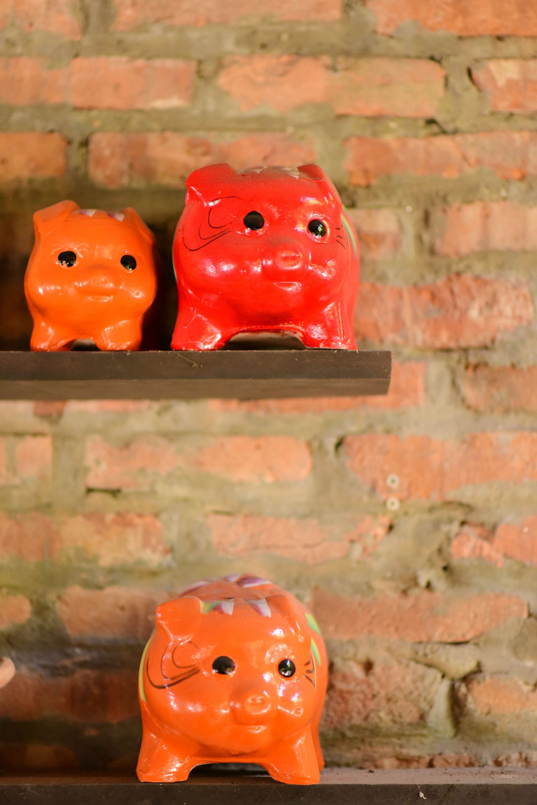 Three vibrant piggy banks on a wooden shelf against a rustic brick wall backdrop.