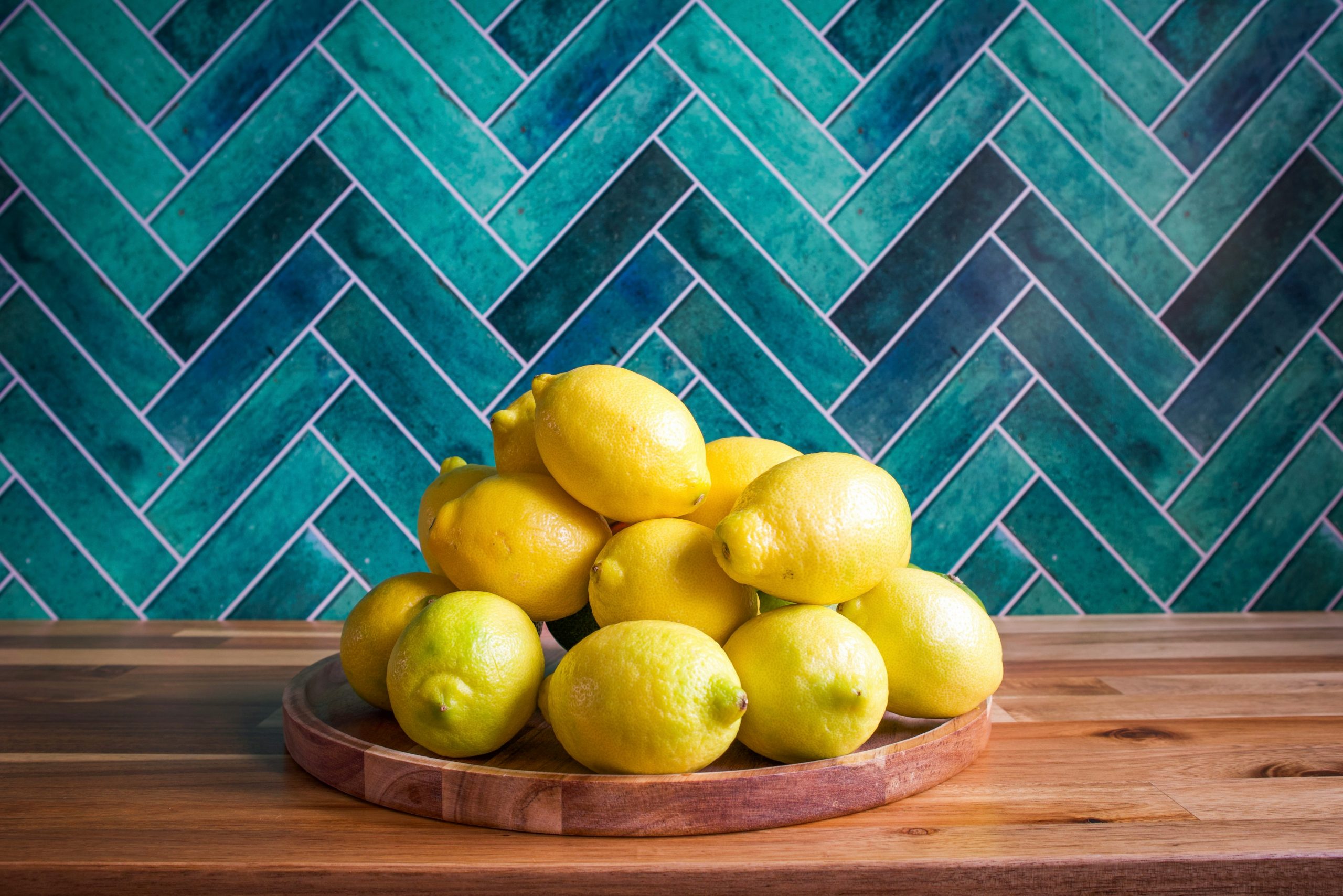 A lively arrangement of fresh yellow lemons sits atop a round wooden tray, placed on a polished wooden countertop. Behind the lemons, a bold teal herringbone tile backsplash adds a modern and artistic