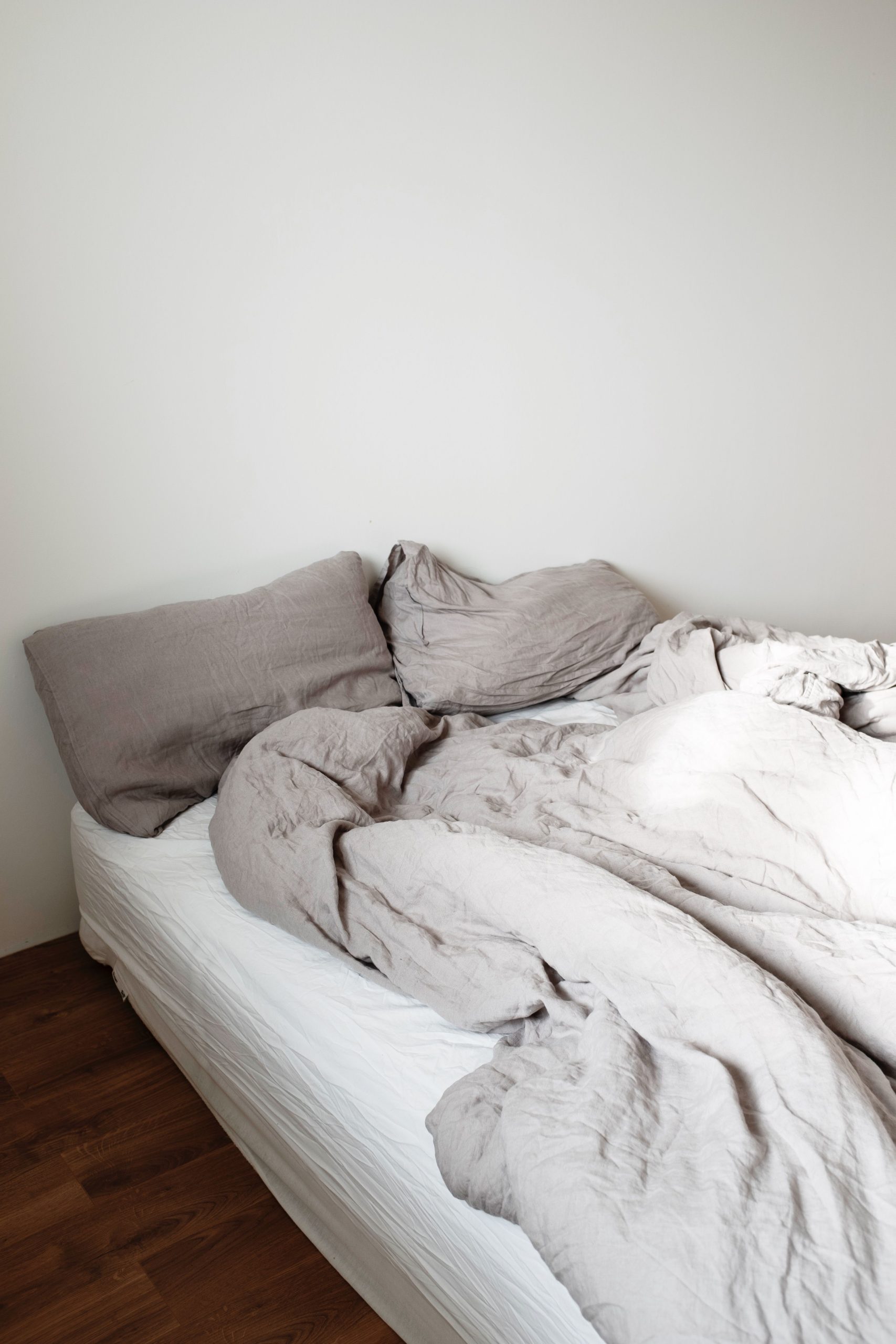 A minimalist bedroom with an unmade bed, featuring soft, wrinkled linen and a wooden floor.