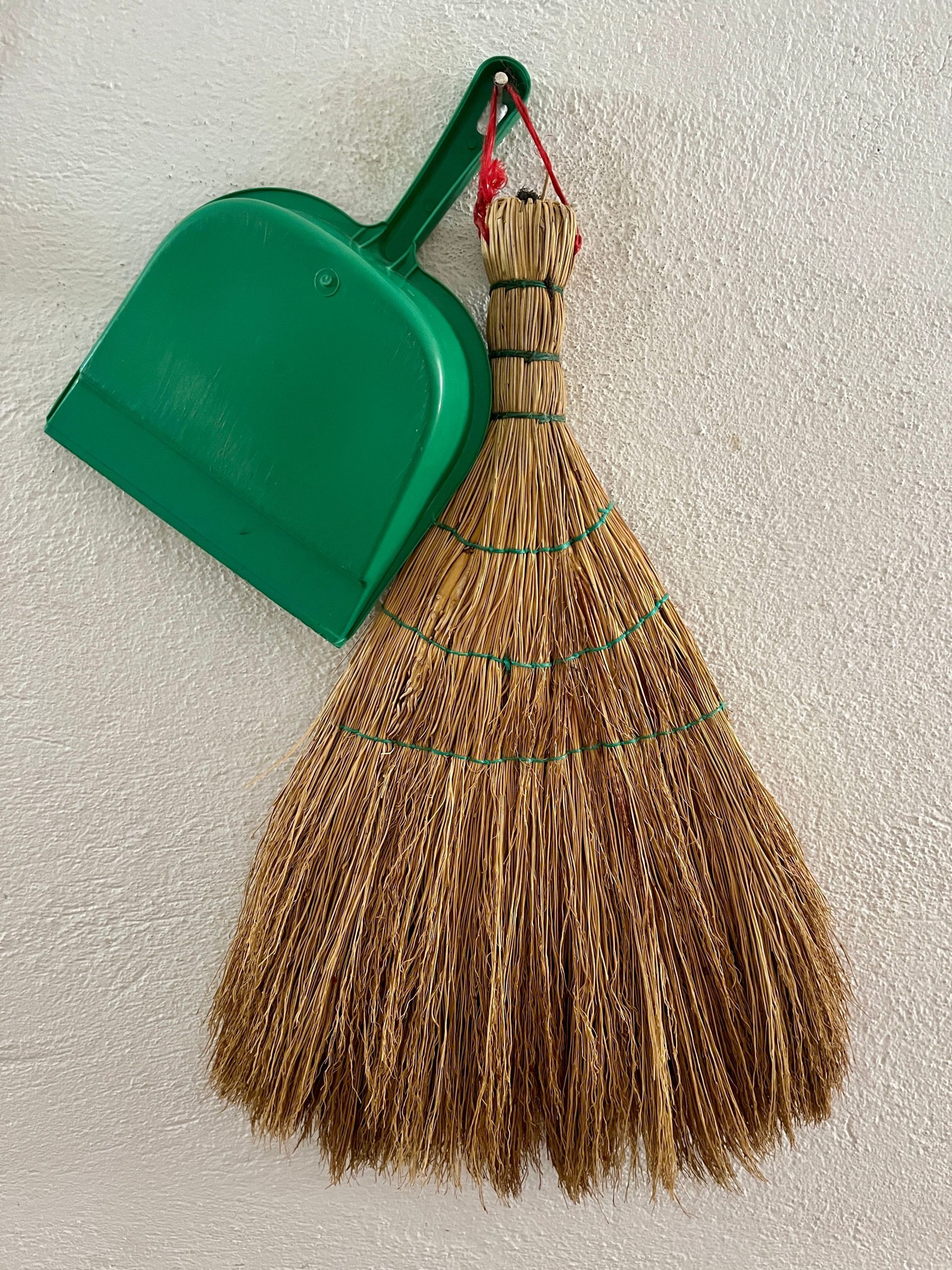 A traditional straw broom and green dustpan hang on a textured wall, symbolizing simple and practical clean living.