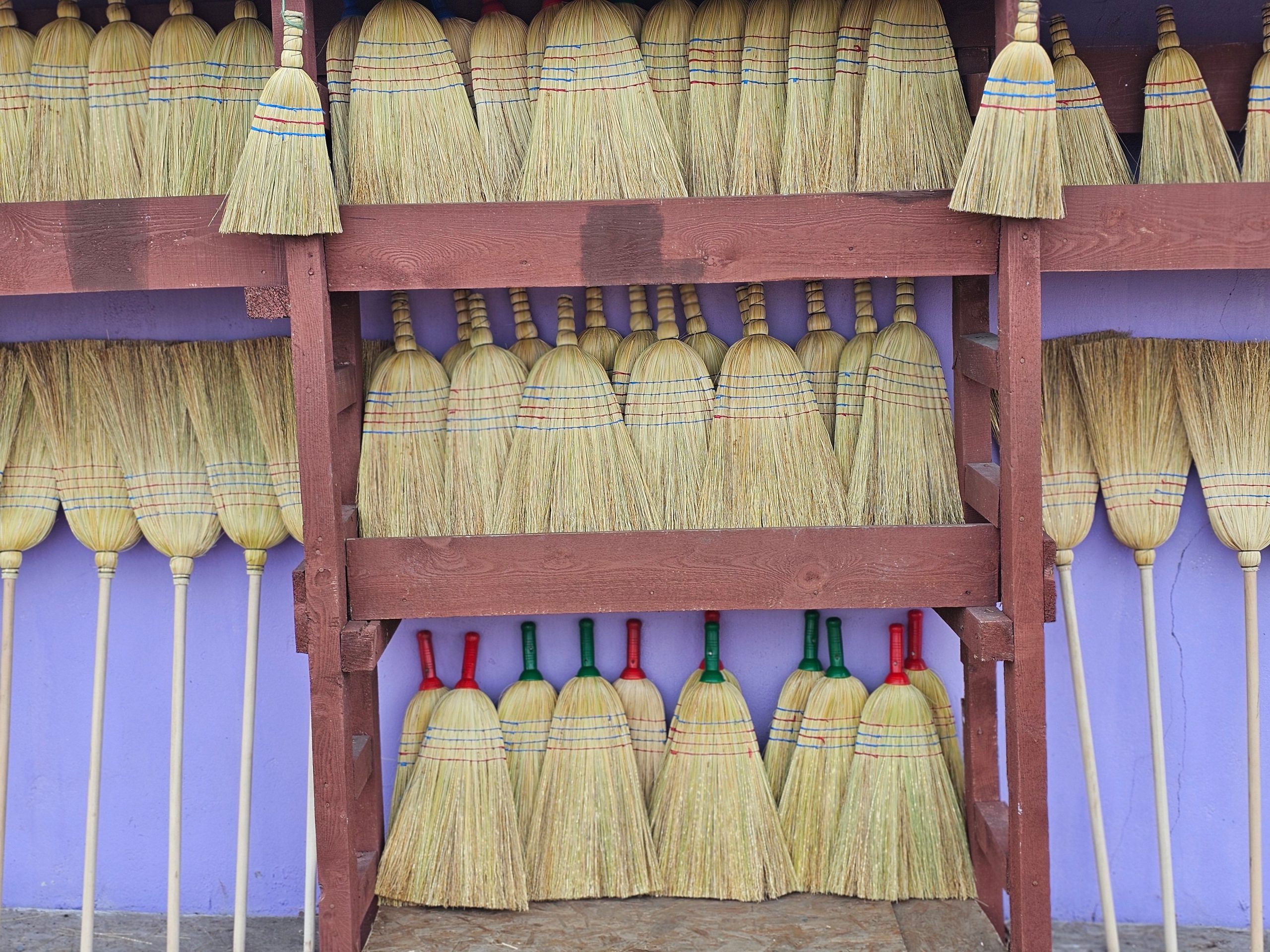 Collection of rustic straw brooms arranged on wooden shelves against a vibrant purple wall.