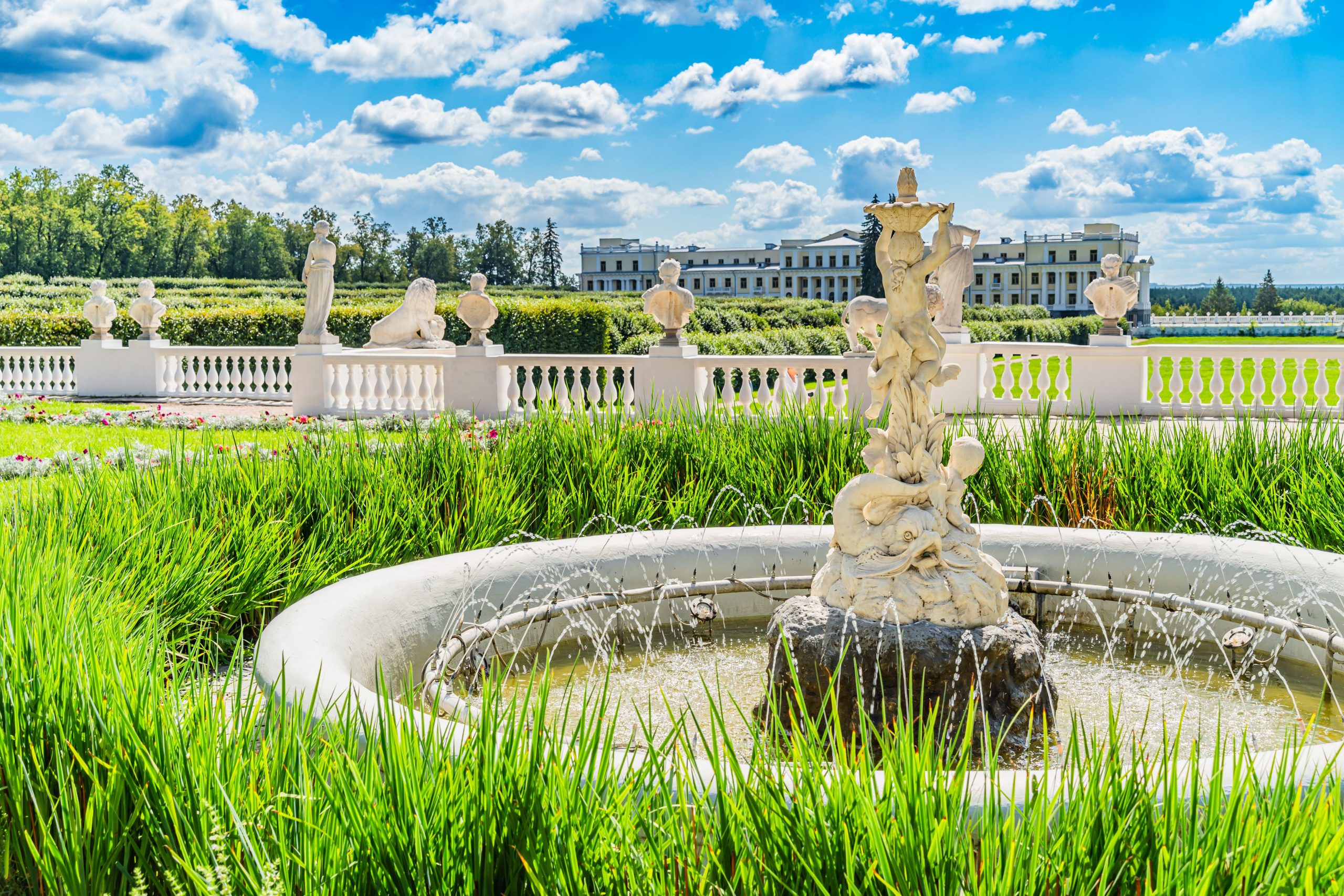 Elegant garden with classical stone fountain and sculptures, set against a historic European building under a bright sky.