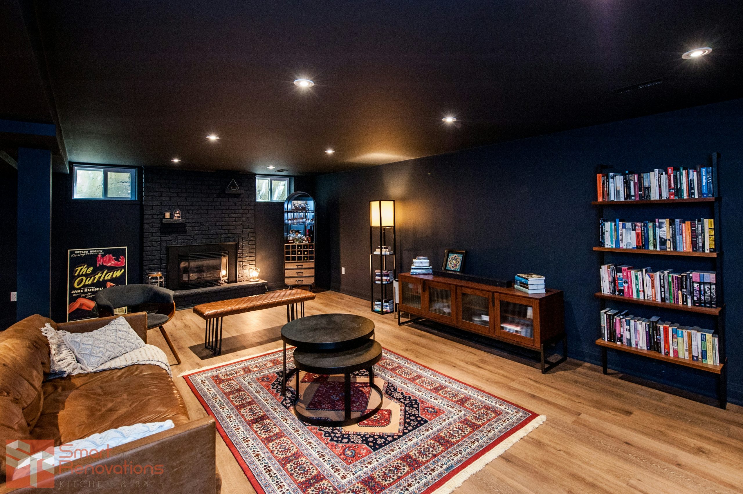 Moody basement lounge styled with deep navy walls, warm oak-look flooring, and a striking black brick fireplace as the focal point. Layered lighting, a rich Persian rug, leather sofa, and full wall of