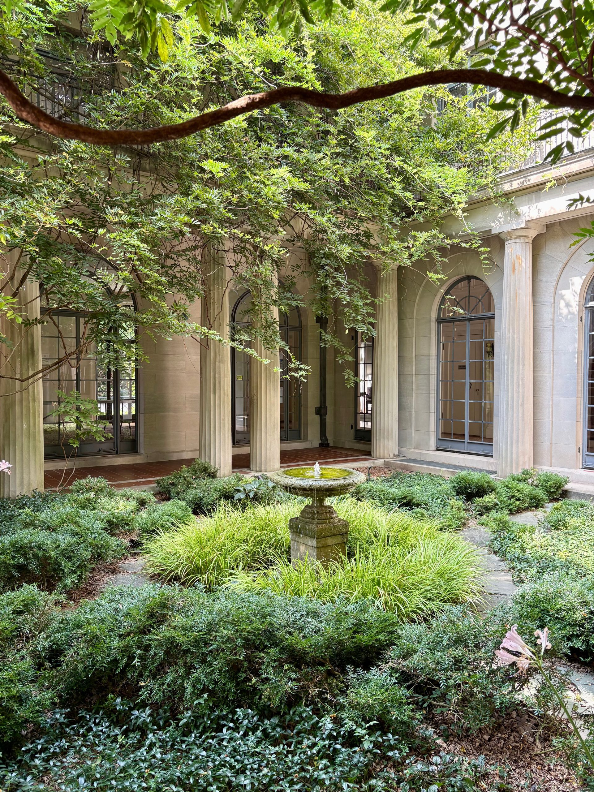 Serene courtyard with lush greenery and stone fountain, framed by classic columns in urban setting.