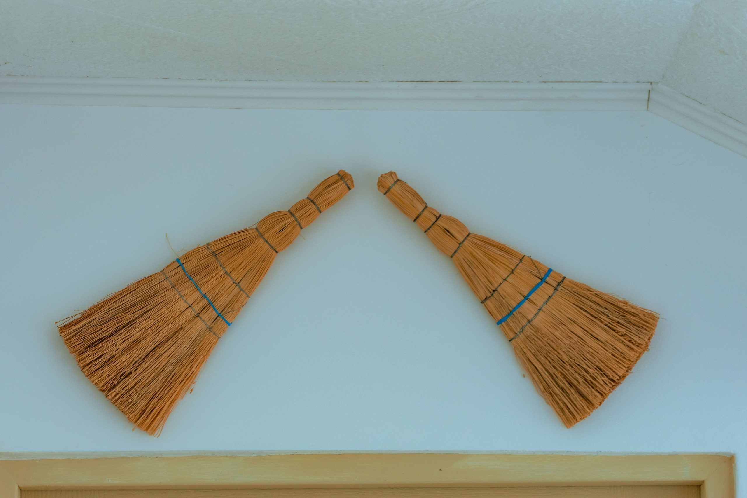 Two decorative rustic straw brooms hanging on a light colored wall.