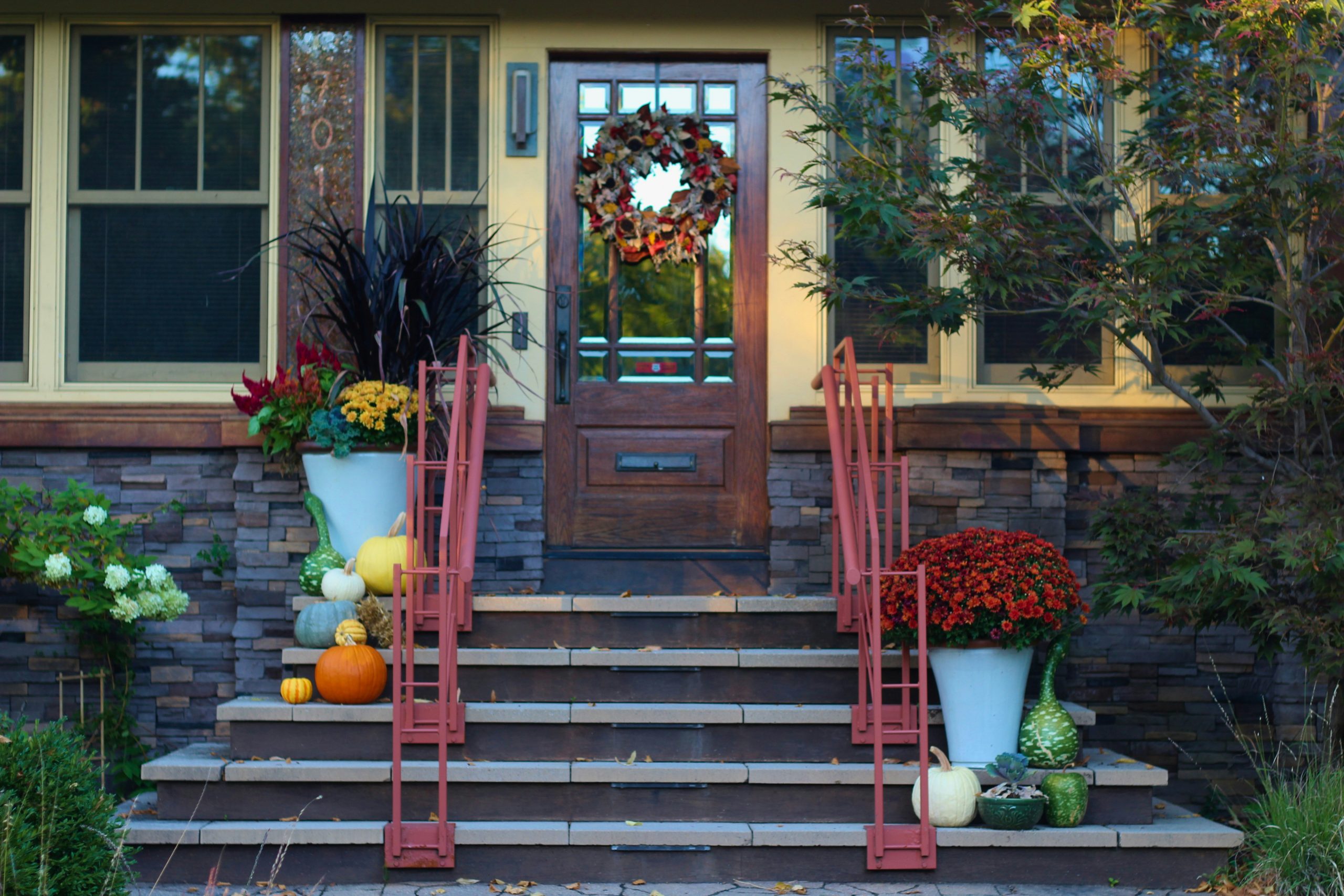 A charming front porch decorated for autumn. The front door is adorned with a fall wreath and flanked by potted mums. The steps are lined with planters filled with pumpkins, gourds, and flowers. The h
