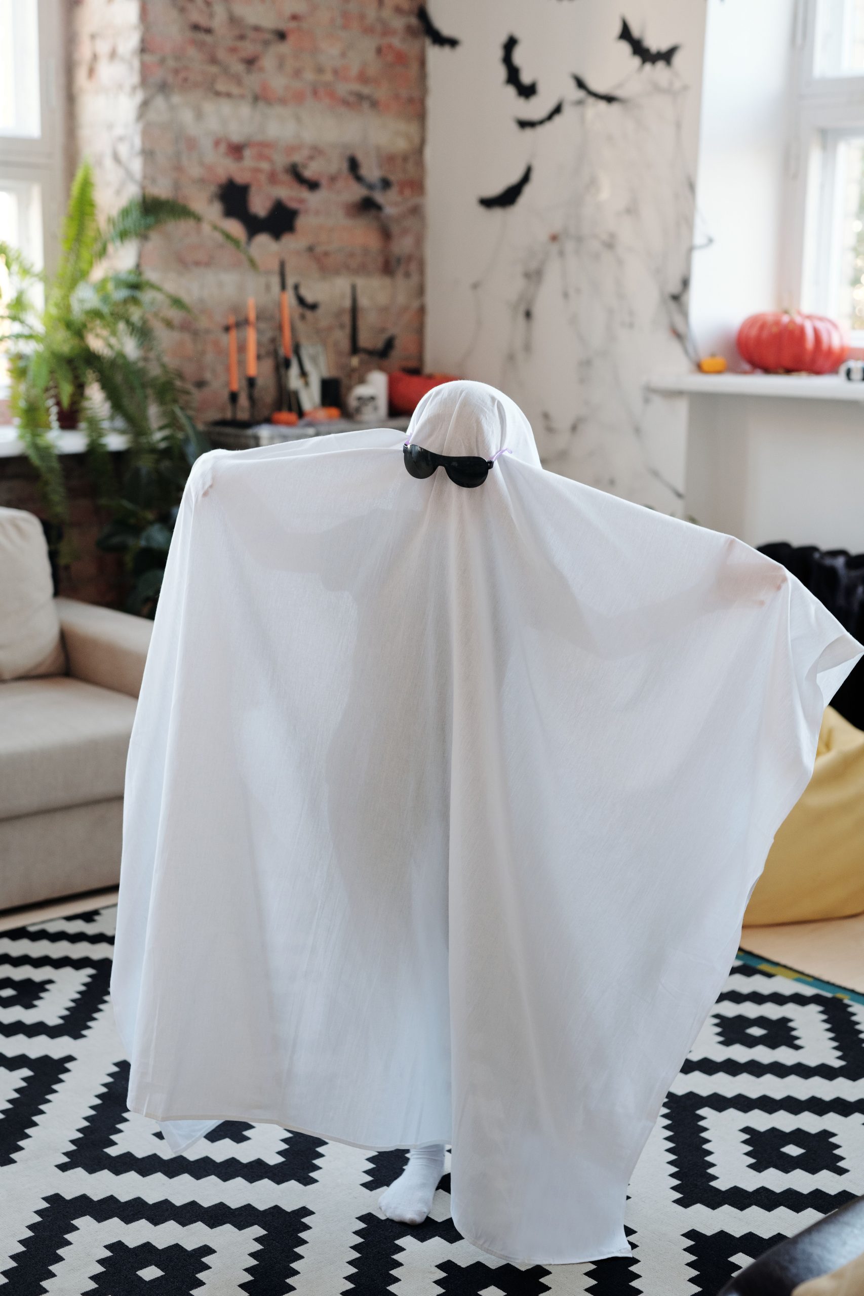 A child dressed as a ghost with sunglasses, enjoying a fun Halloween setup indoors.