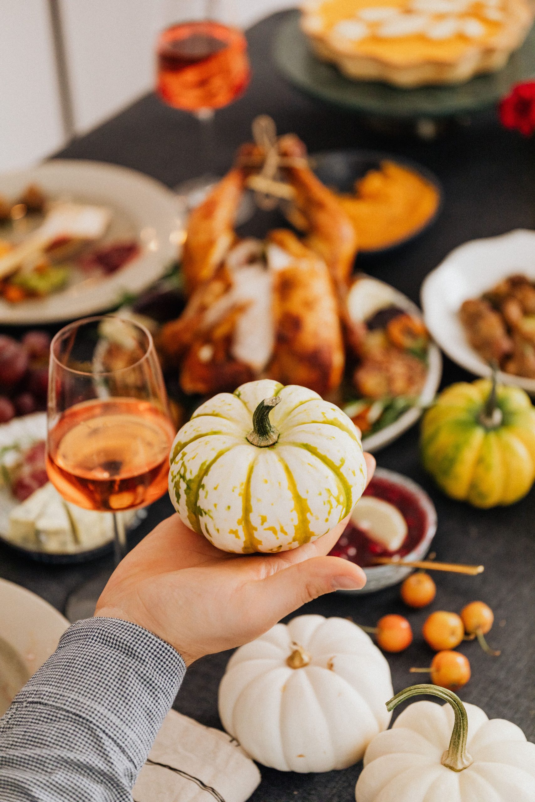 A hand holding a small ornamental pumpkin over a fall-themed dinner table with roasted turkey and wine.