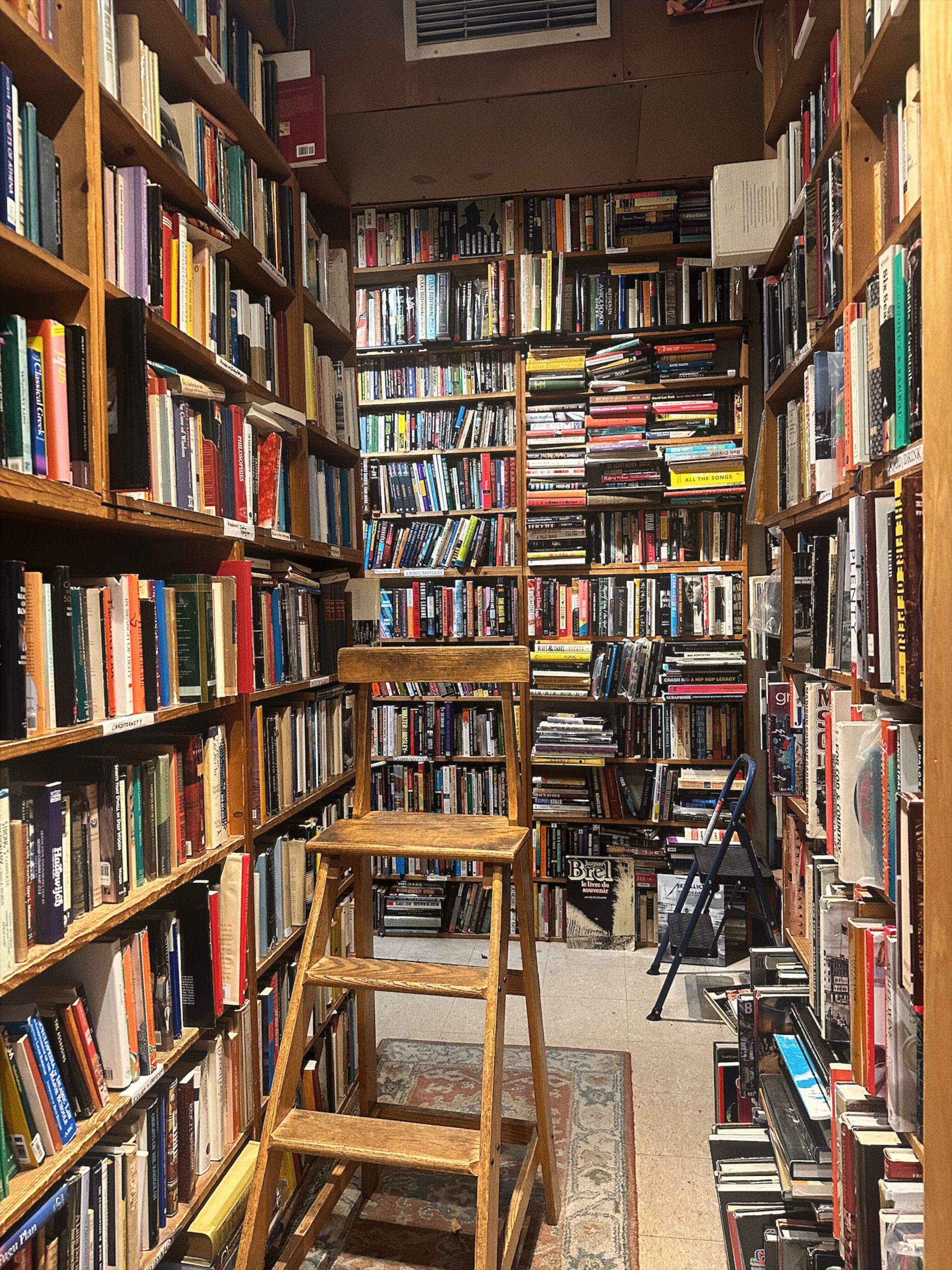 A narrow library aisle filled with bookshelves, featuring a wooden ladder for access.