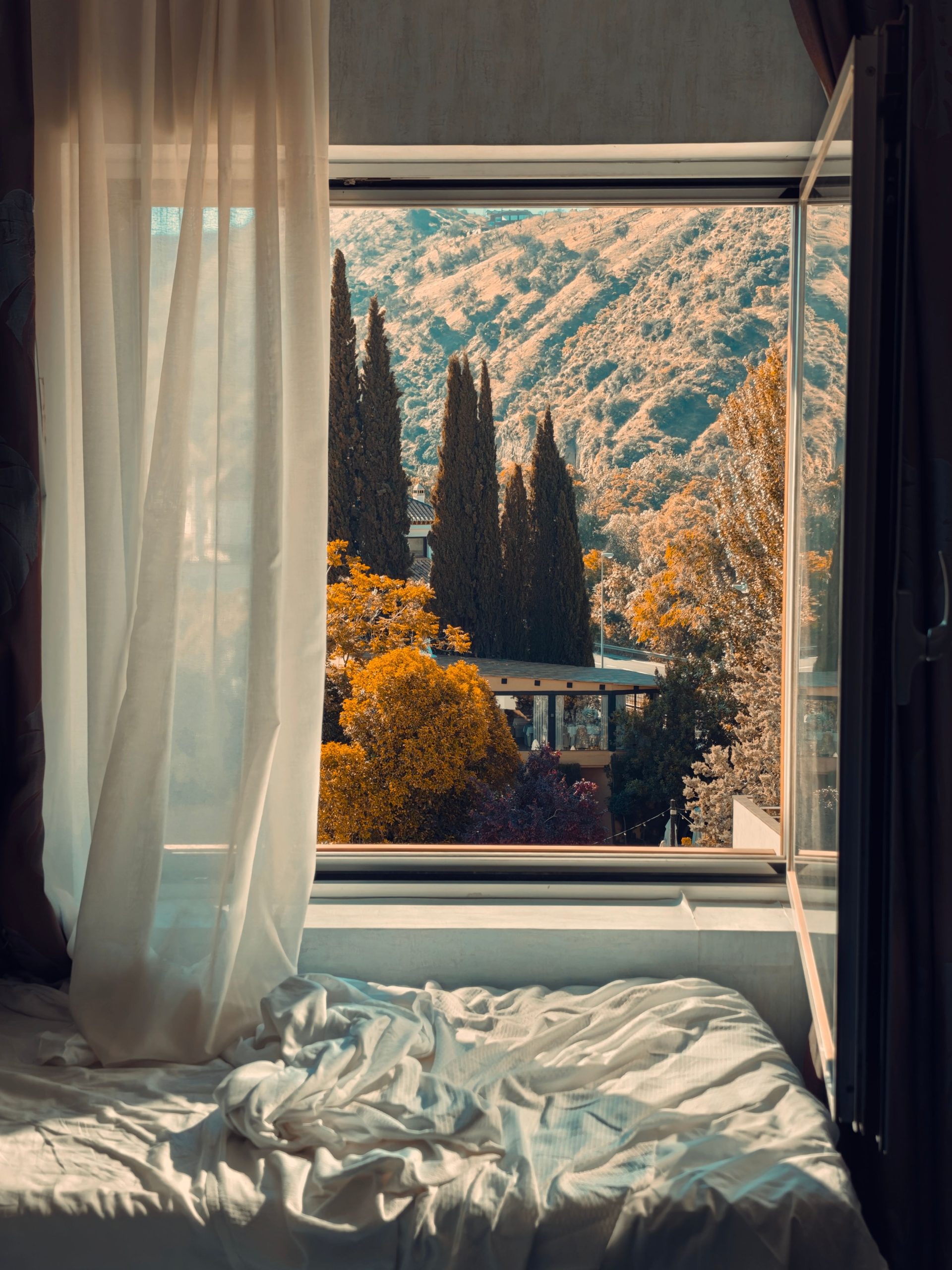 A serene window view from a cozy bedroom overlooking a vibrant autumn scenery with hills and trees.