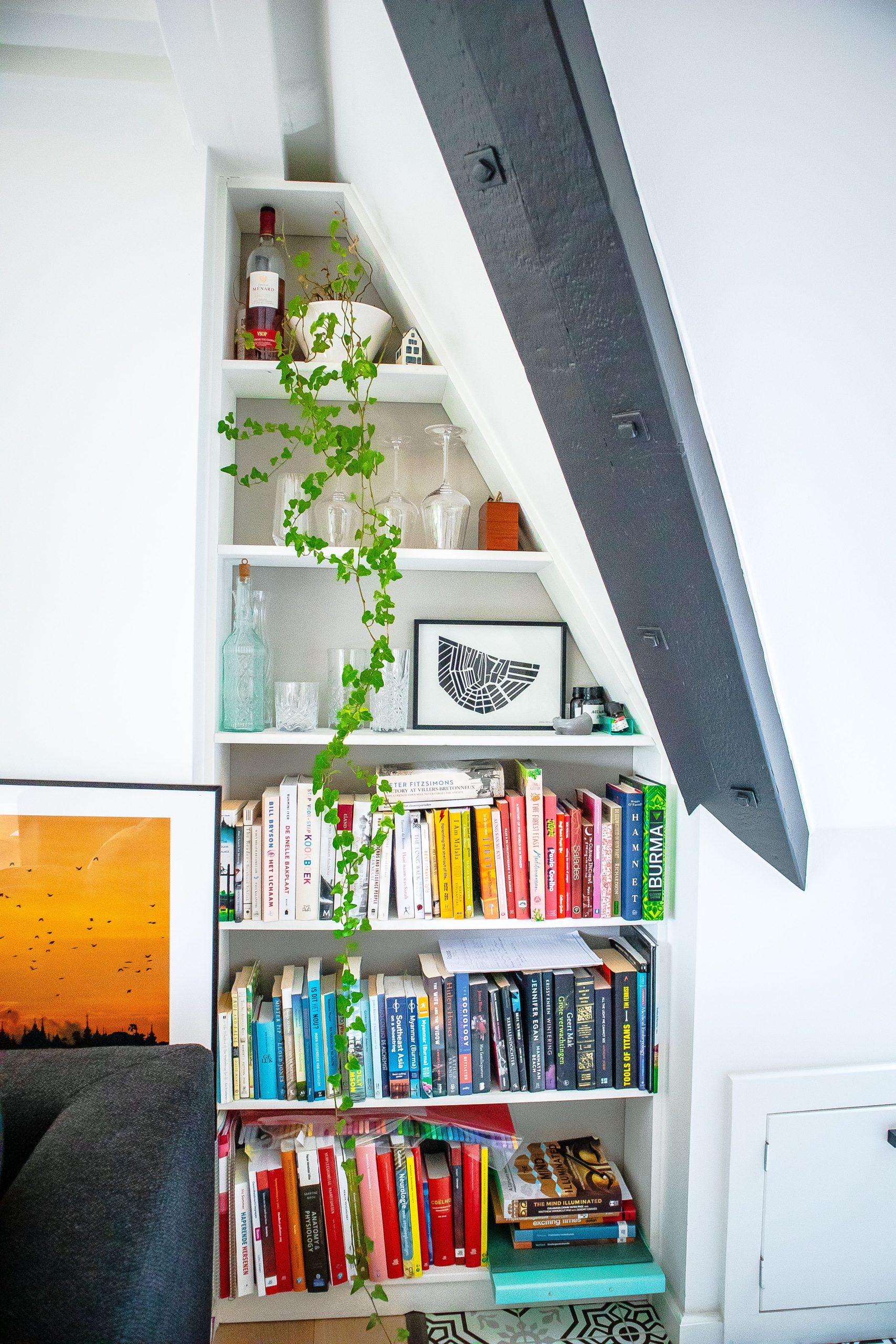 A stylish bookshelf with plants and books in a modern room under stairs.